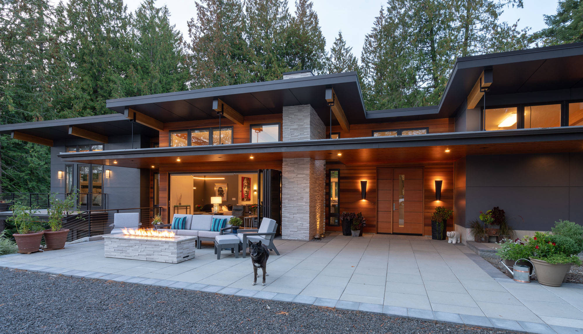 Modern home exterior with cedar siding, stone firepit, concrete patio, and floor-to-ceiling windows backed by evergreen forest.