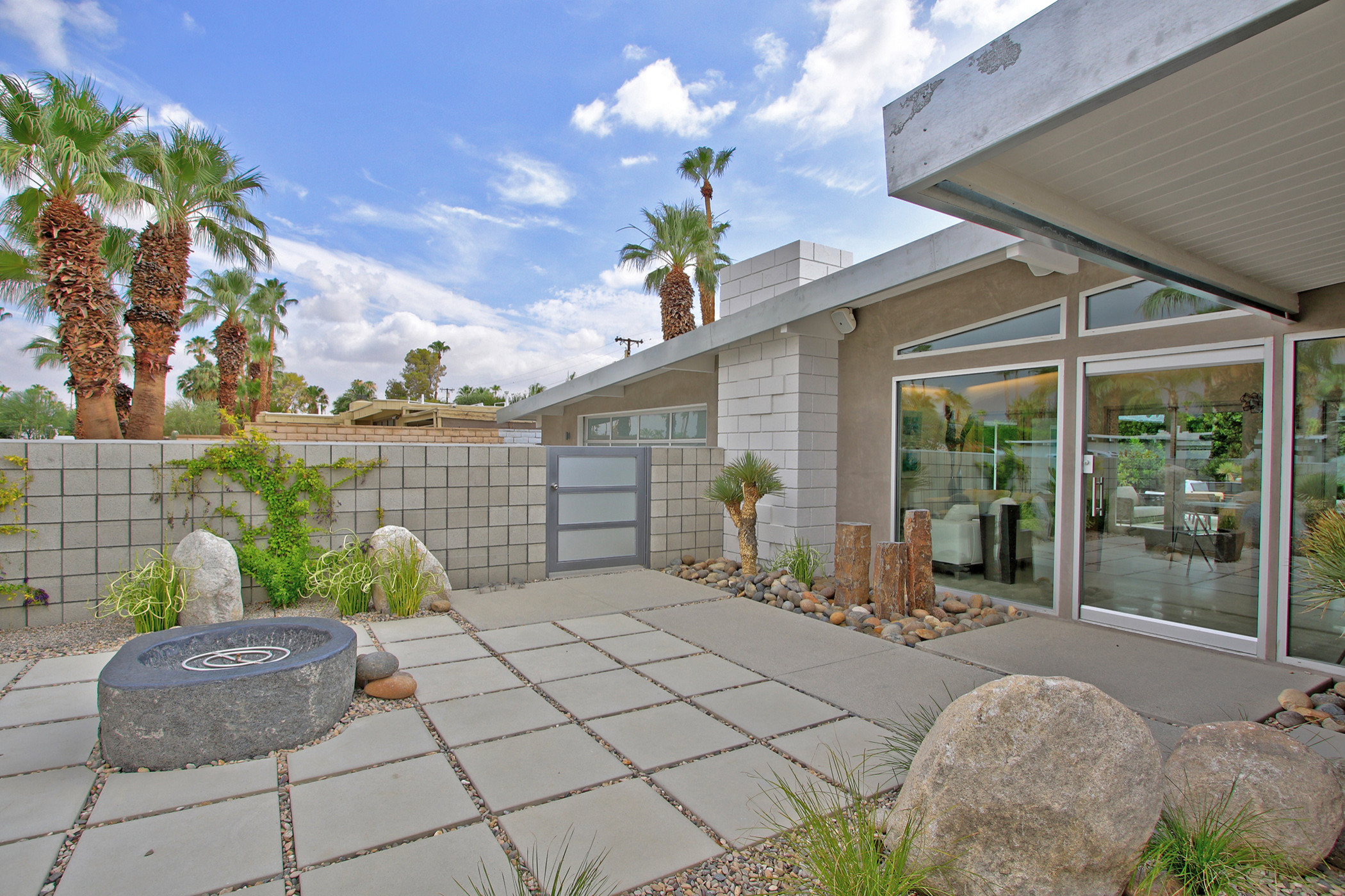 Modern desert courtyard featuring concrete pavers, boulder accents, palm trees, and floor-to-ceiling windows against white masonry walls.