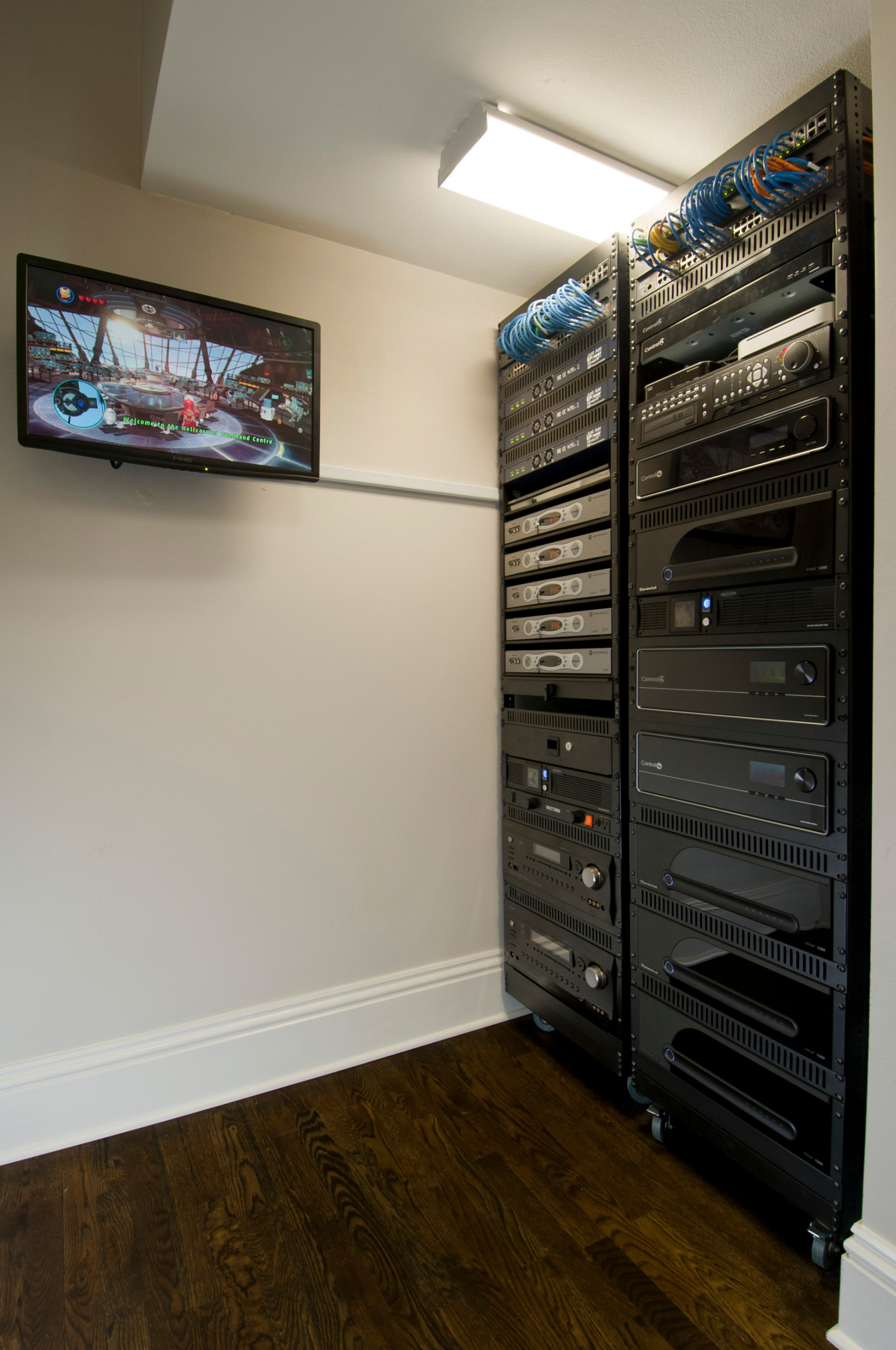 Server room with tall equipment rack, blue cables, wall-mounted display screen, dark wood flooring, and white walls.