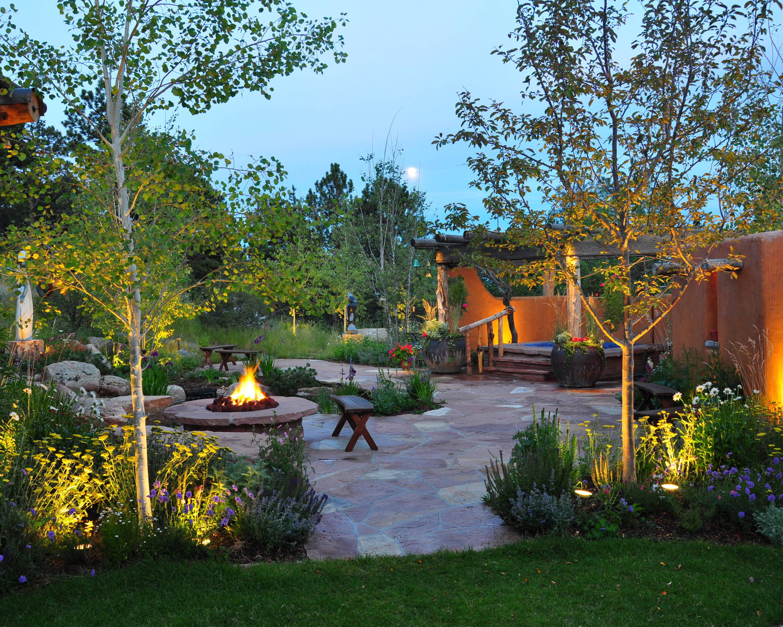 Evening garden courtyard featuring circular firepit, flagstone patio, illuminated birch trees, and adobe walls with native plantings.