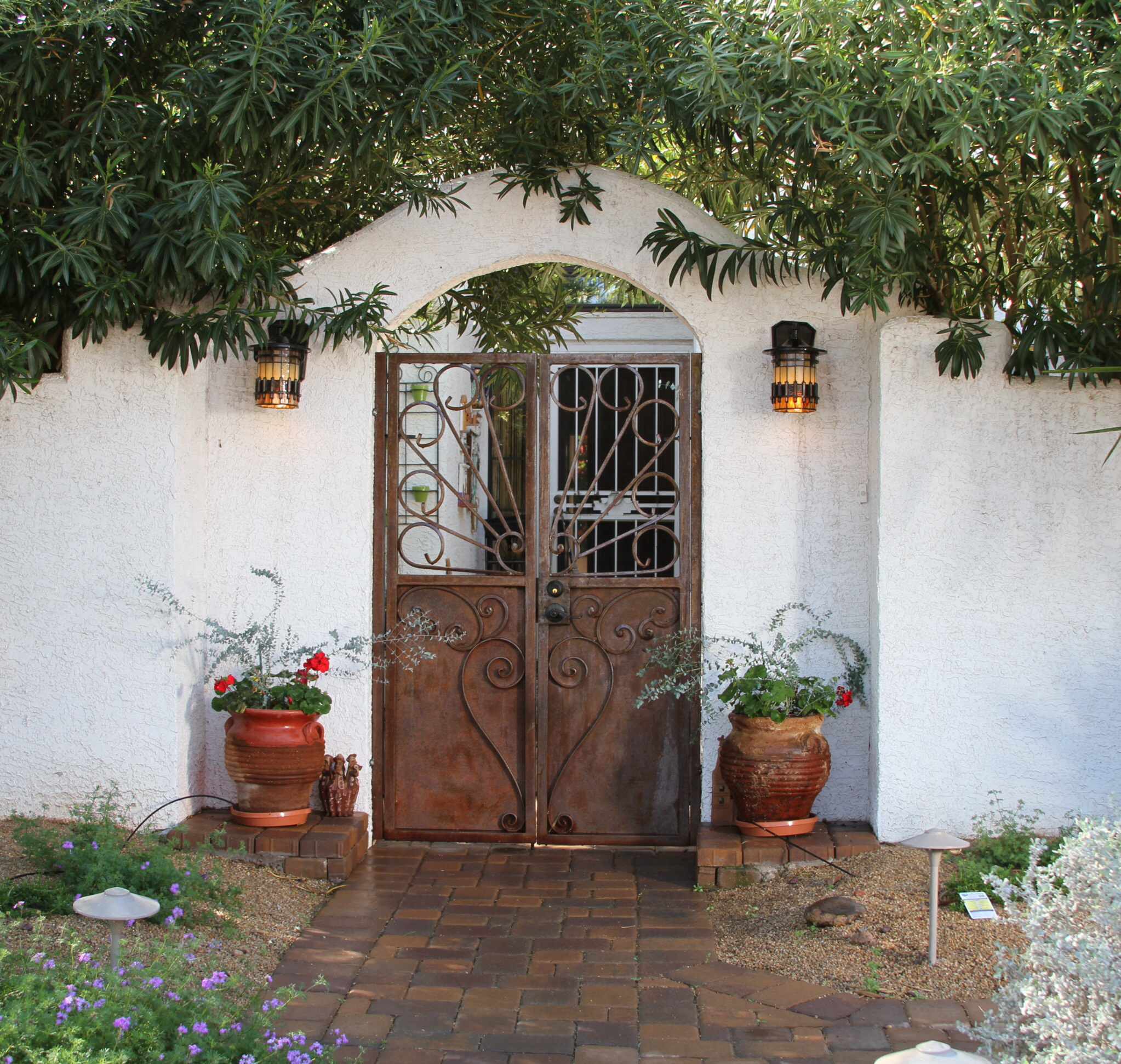 Arched entryway with ornate iron double doors, flanked by terra cotta planters and amber lanterns under leafy canopy.