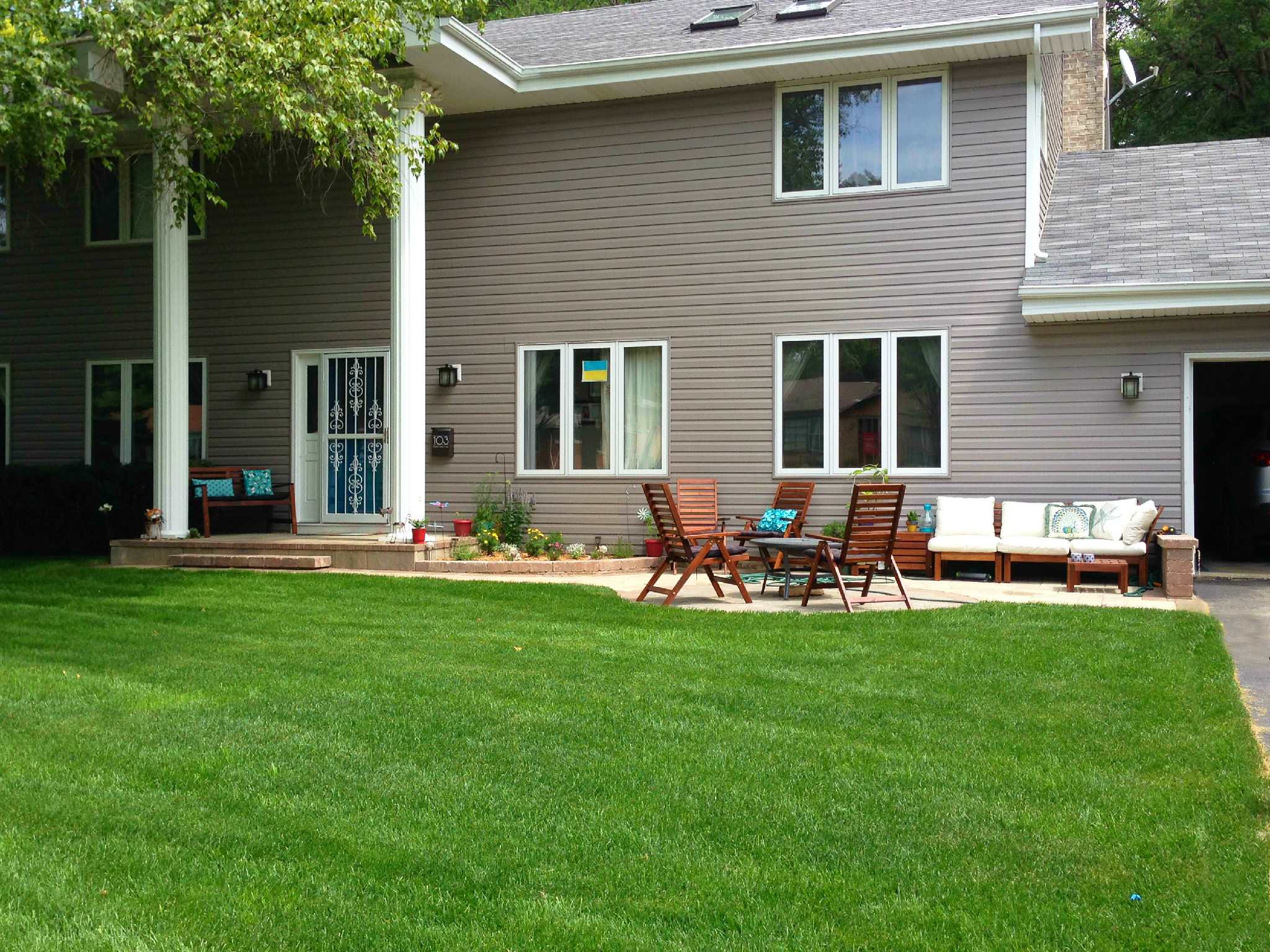 Concrete patio with teak wooden chairs and bench, turquoise accent pillows, bordered by manicured lawn and taupe house exterior.
