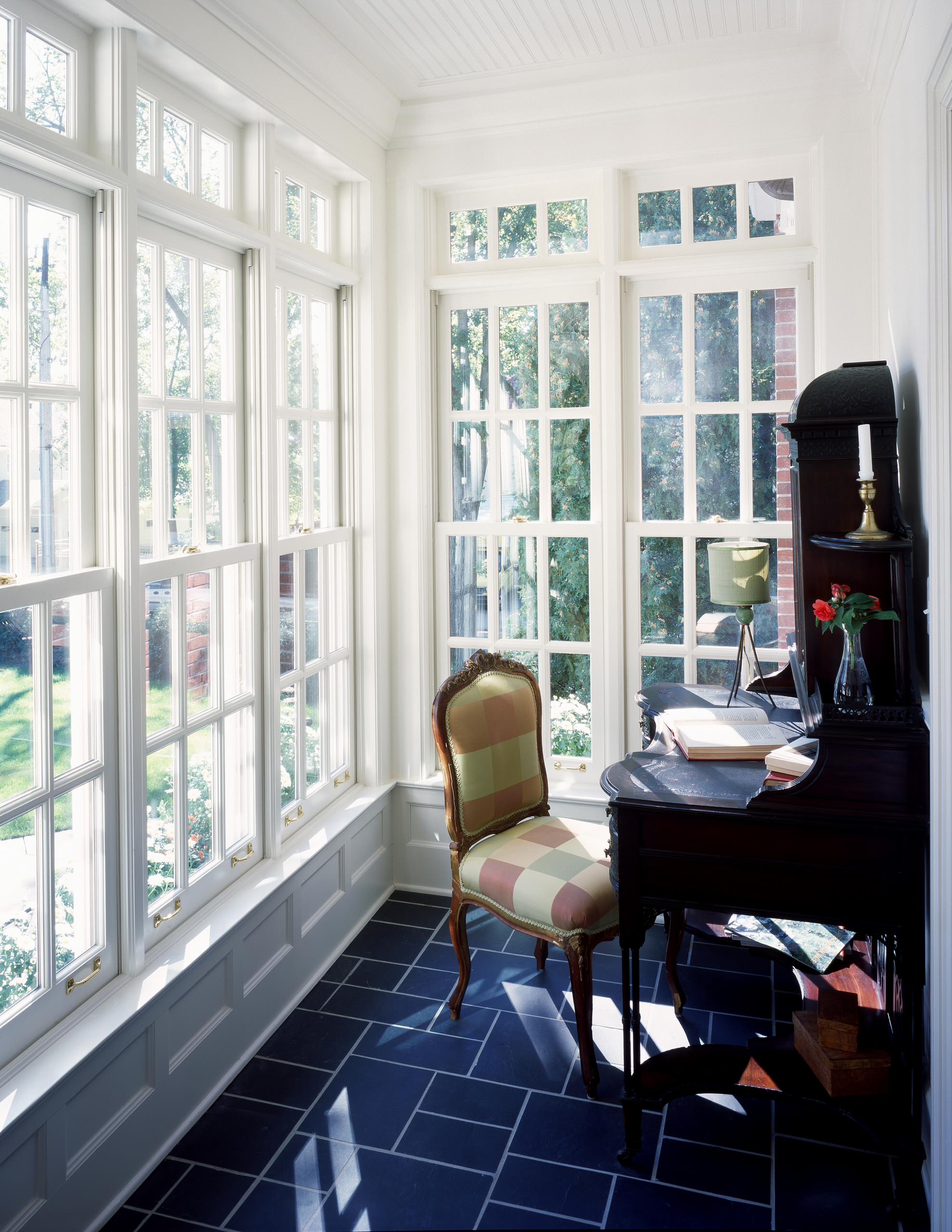 Bright sunroom with black piano, checkered upholstered chair, slate flooring, and floor-to-ceiling French windows overlooking garden.