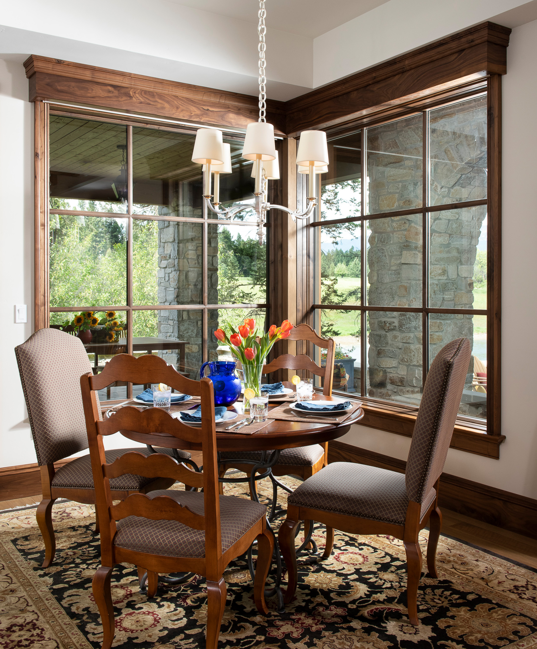 Corner breakfast nook with walnut-trimmed windows, round table, upholstered chairs, and chandelier overlooking mountain landscape.