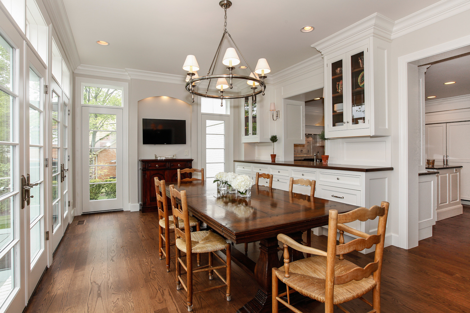 Bright dining room with wall of French doors, mahogany table, rush-seat chairs, circular chandelier, and white built-in cabinetry.