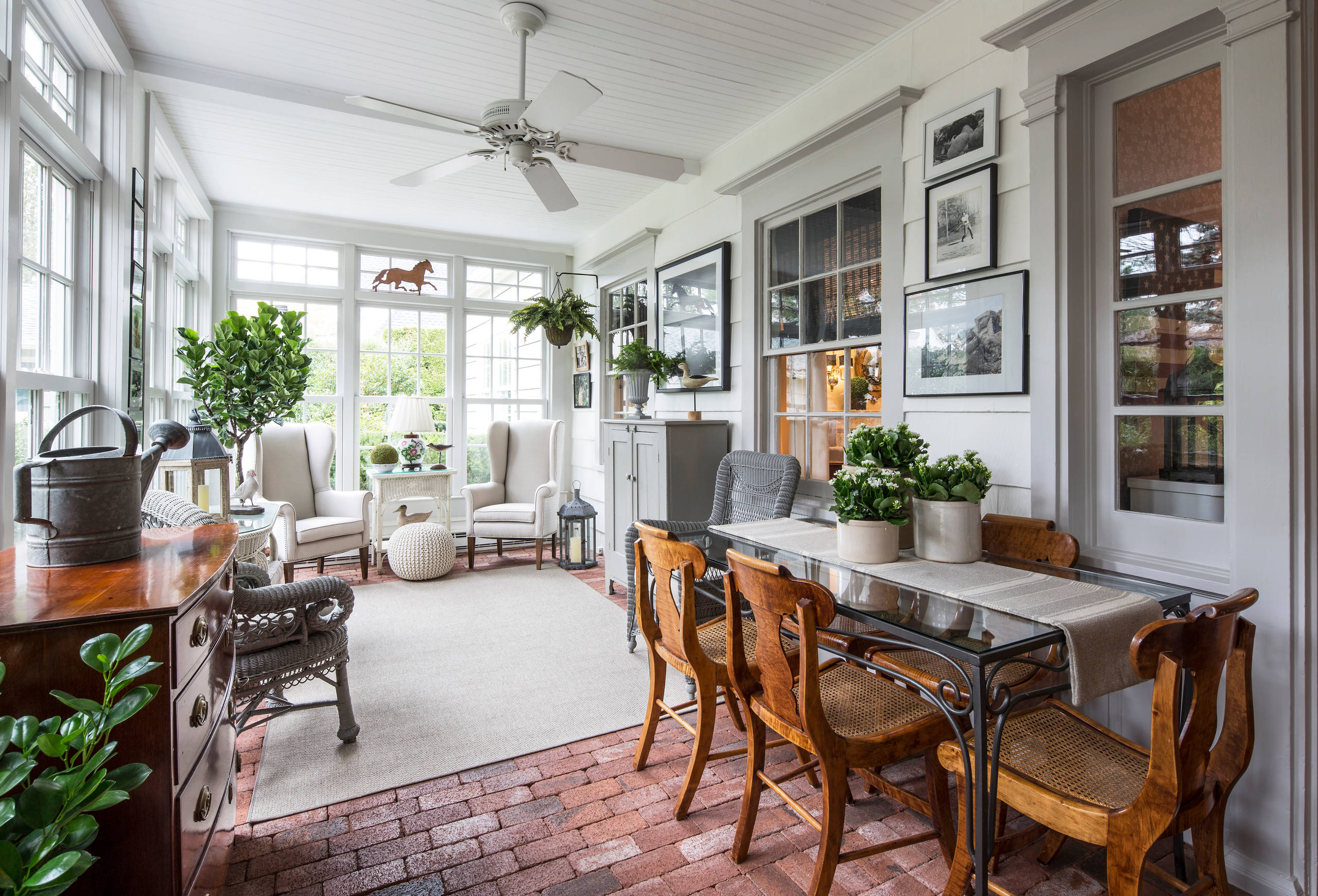 Sunlit enclosed porch with brick flooring, vintage furniture, white wingback chairs, and equestrian artwork featuring abundant greenery.
