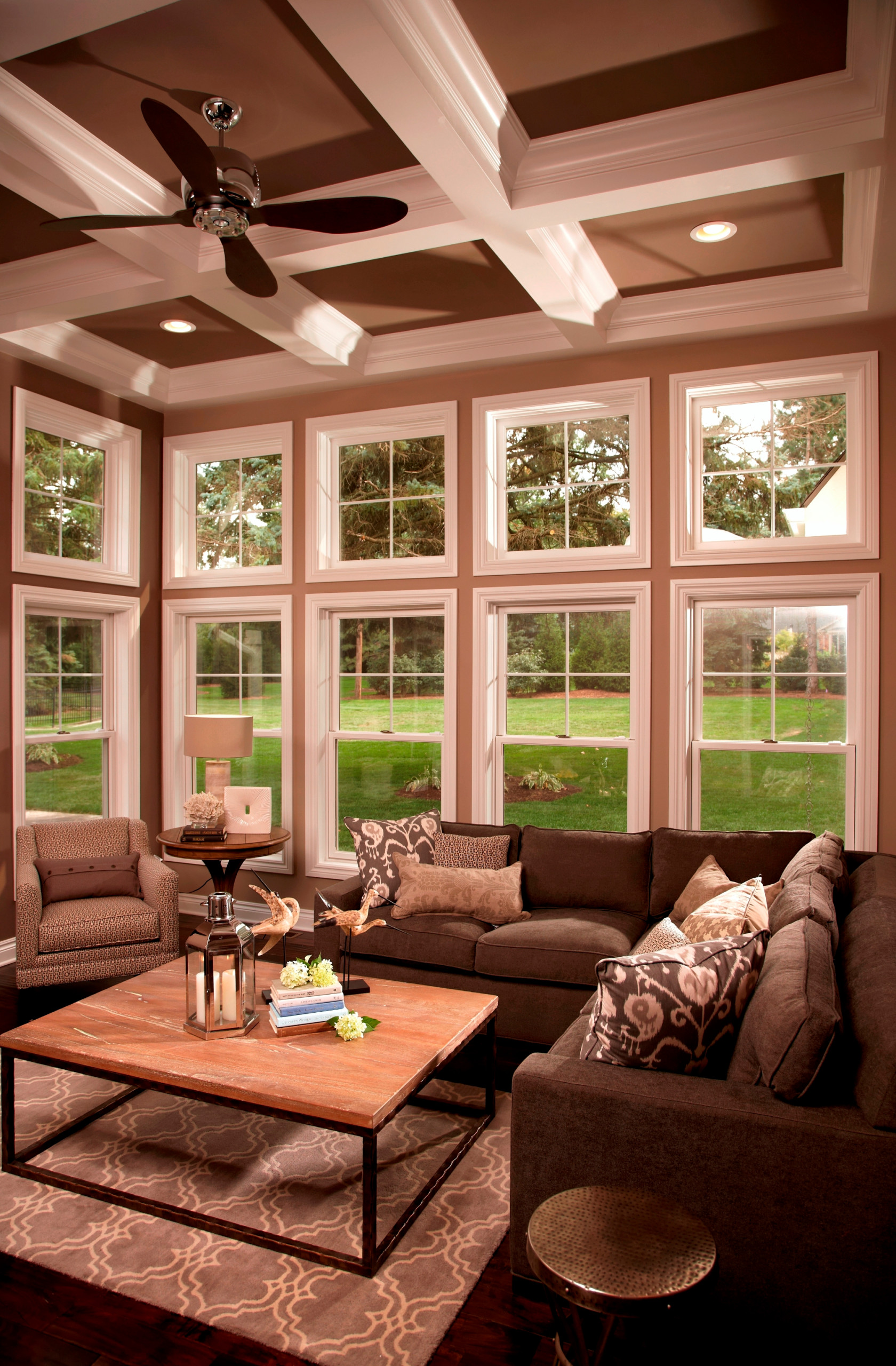 Sophisticated sunroom featuring double-height windows, brown coffered ceiling, sectional sofa, and geometric rug beneath industrial coffee table.