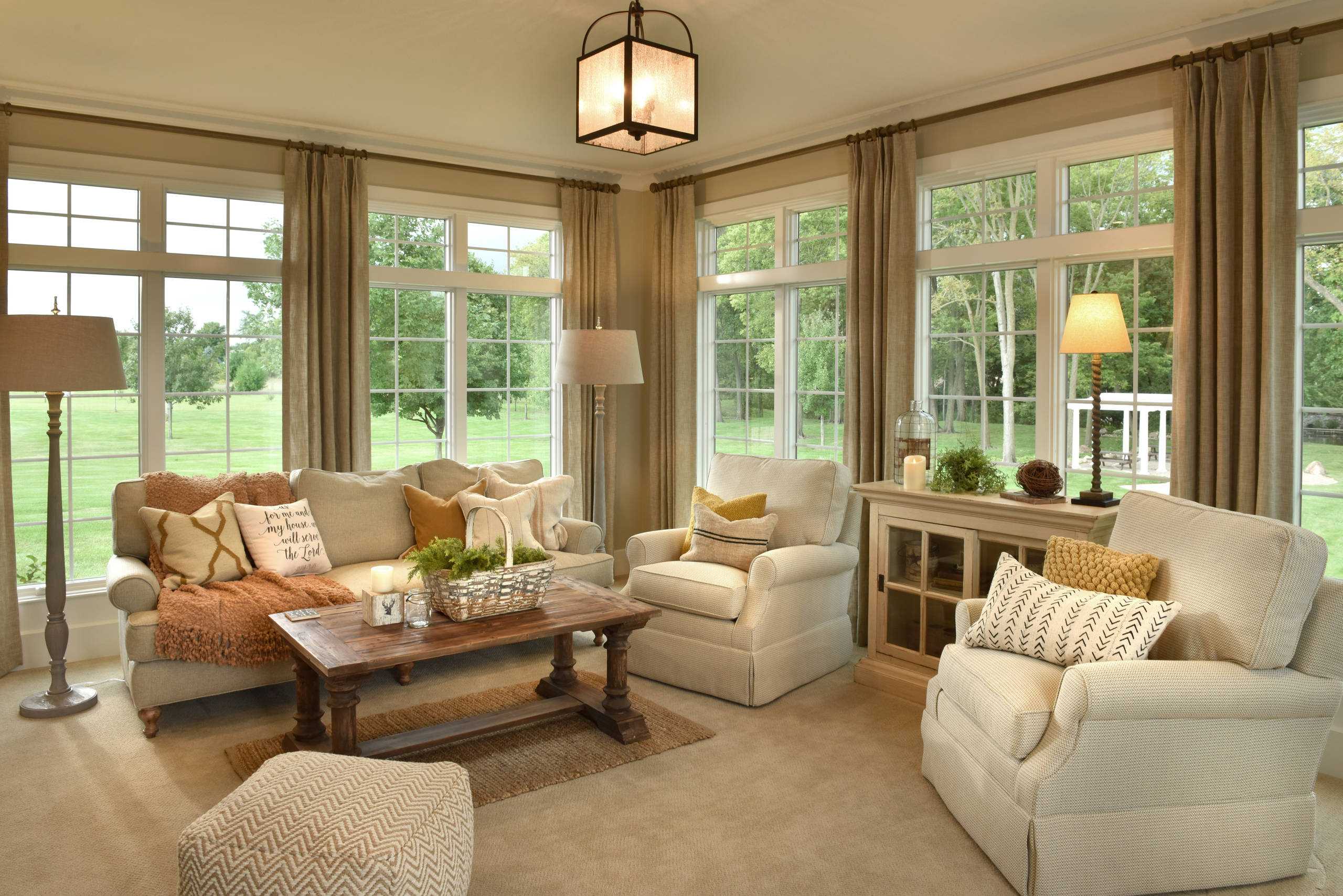 Bright sunroom with cream upholstered furniture, floor-to-ceiling windows, rustic coffee table, and taupe curtains overlooking pastoral landscape.