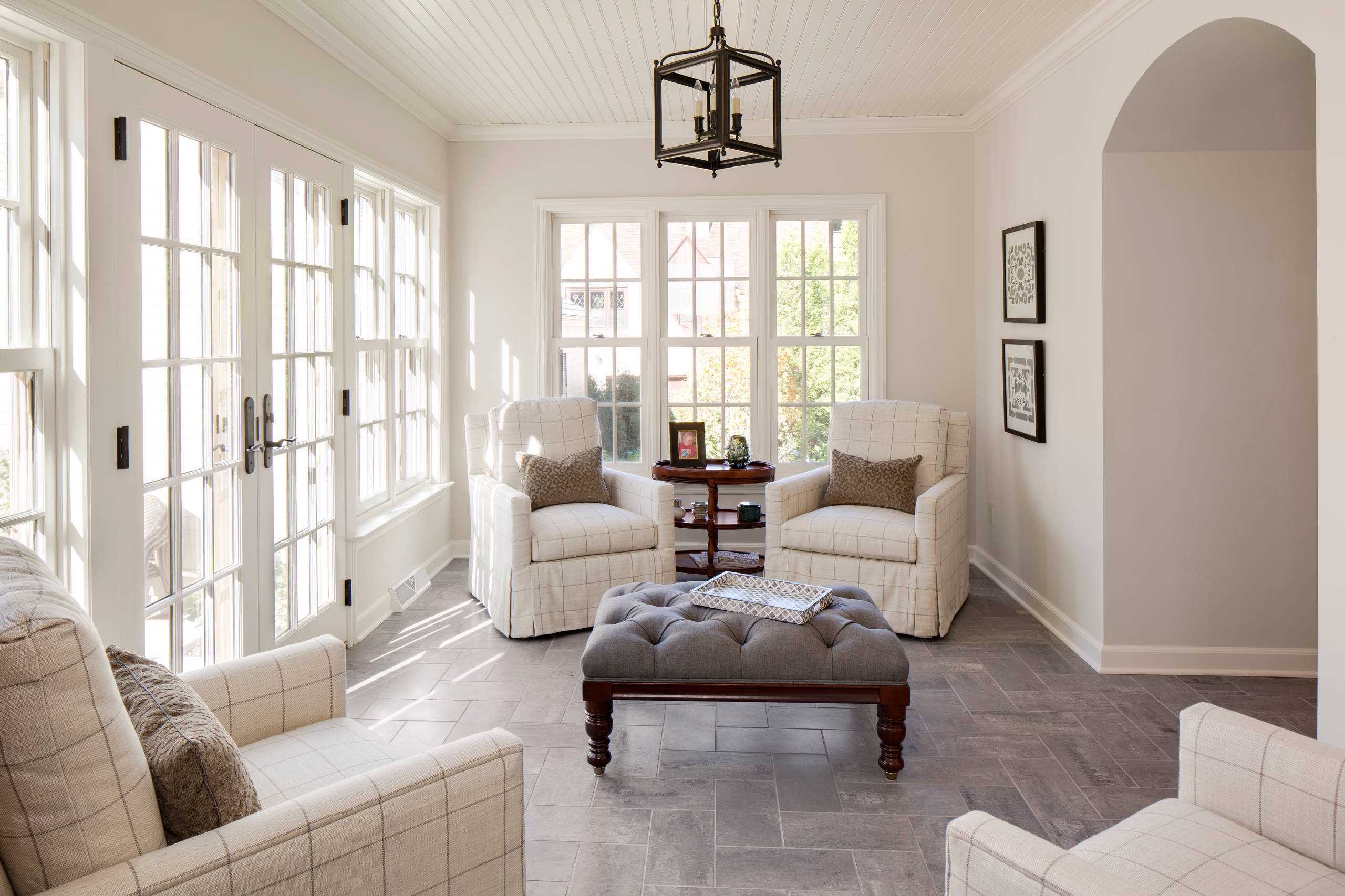 Bright sunroom featuring windowpane-upholstered chairs, gray herringbone tile floor, and floor-to-ceiling French windows.