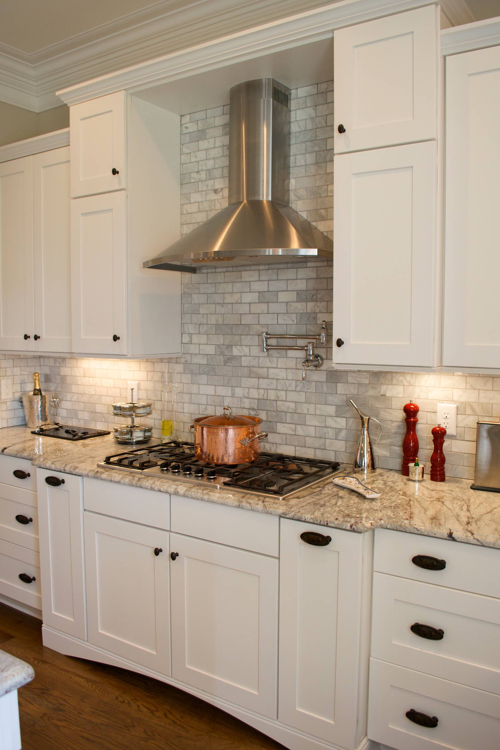 White kitchen featuring marble subway tile backsplash, stainless range hood, granite countertops, and copper cookware on gas cooktop.