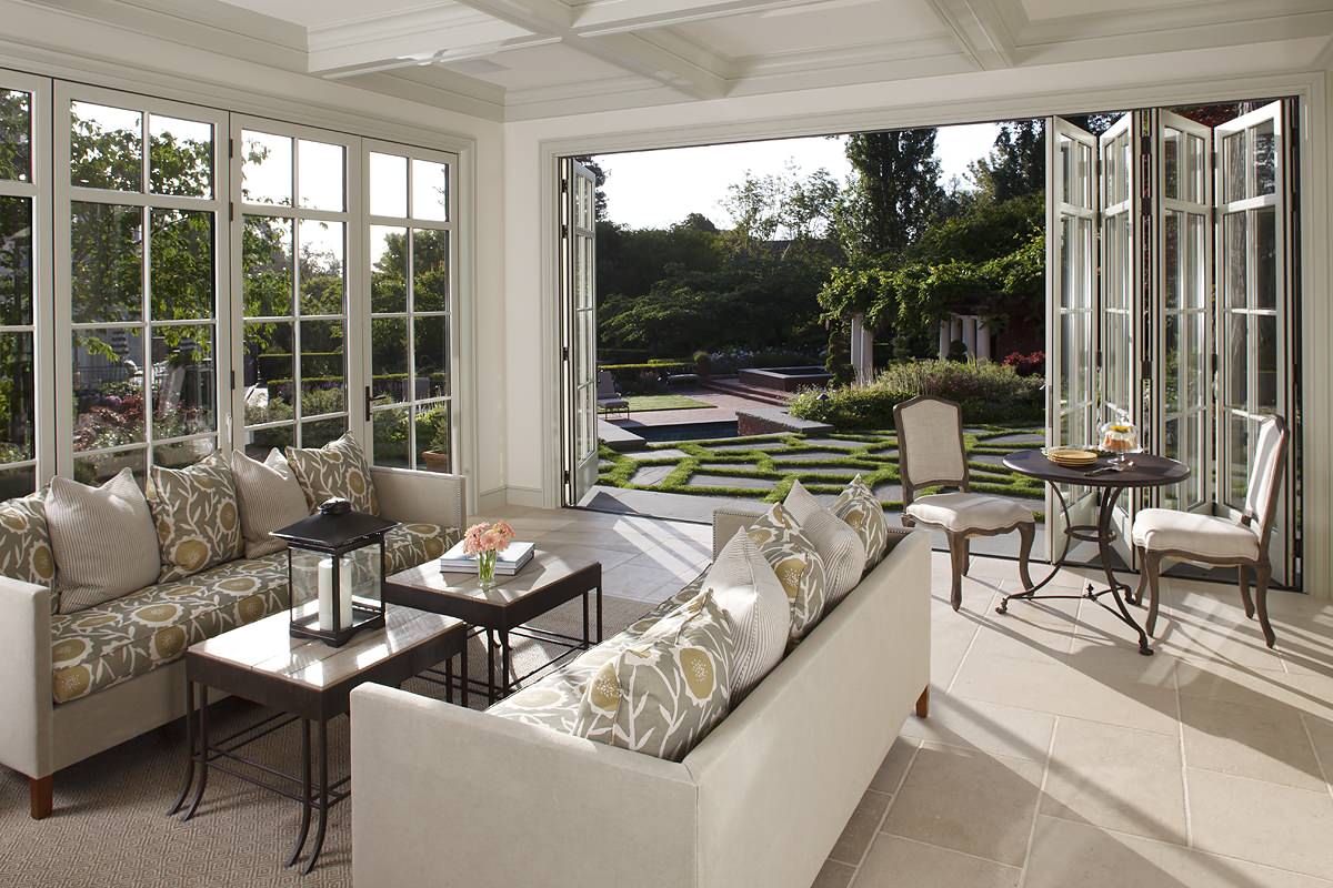 Bright sunroom with white coffered ceiling, folding glass doors, cream upholstered seating, and geometric garden views.