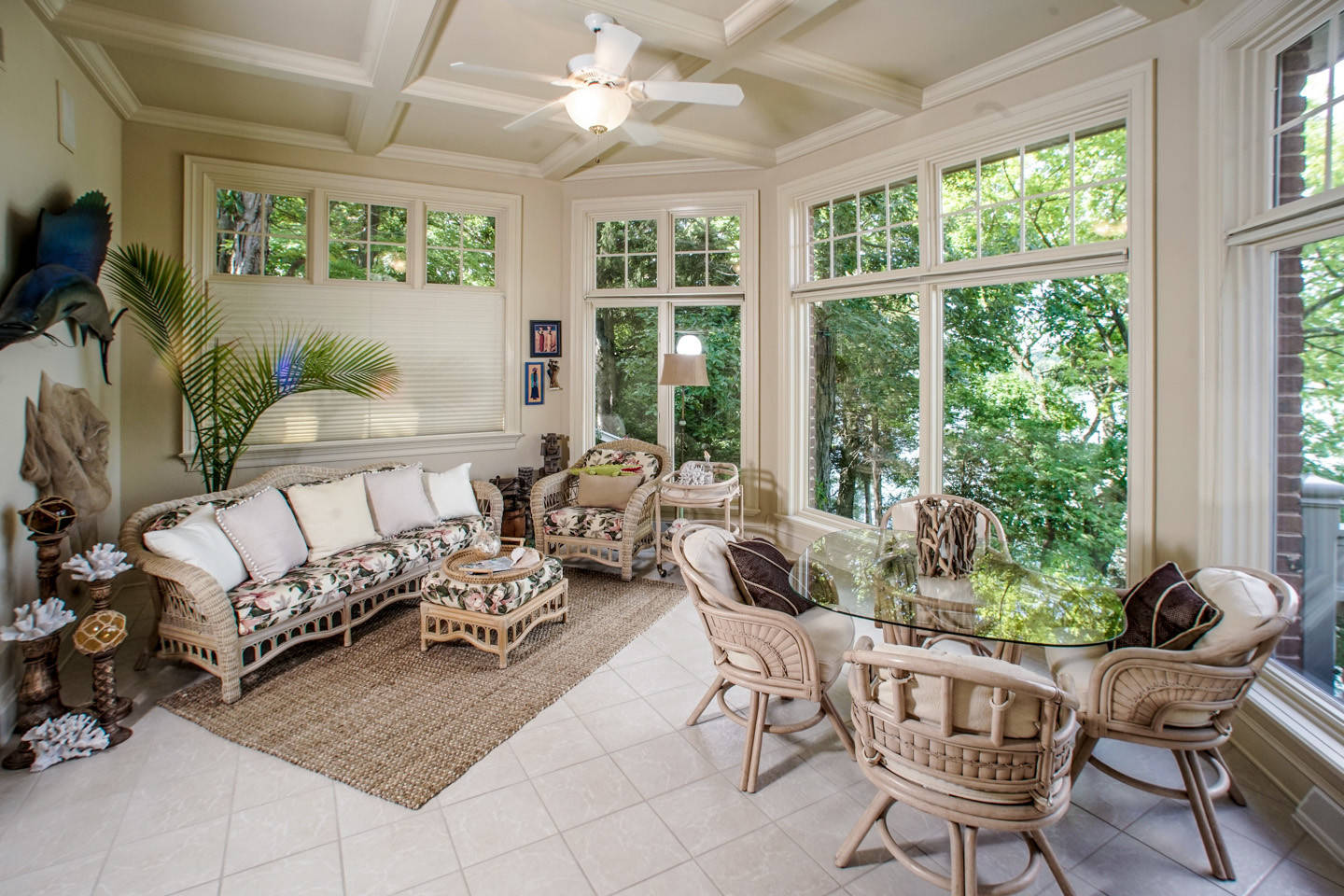Bright sunroom with white coffered ceiling, wicker furniture set, floral cushions, and large windows overlooking lush greenery.