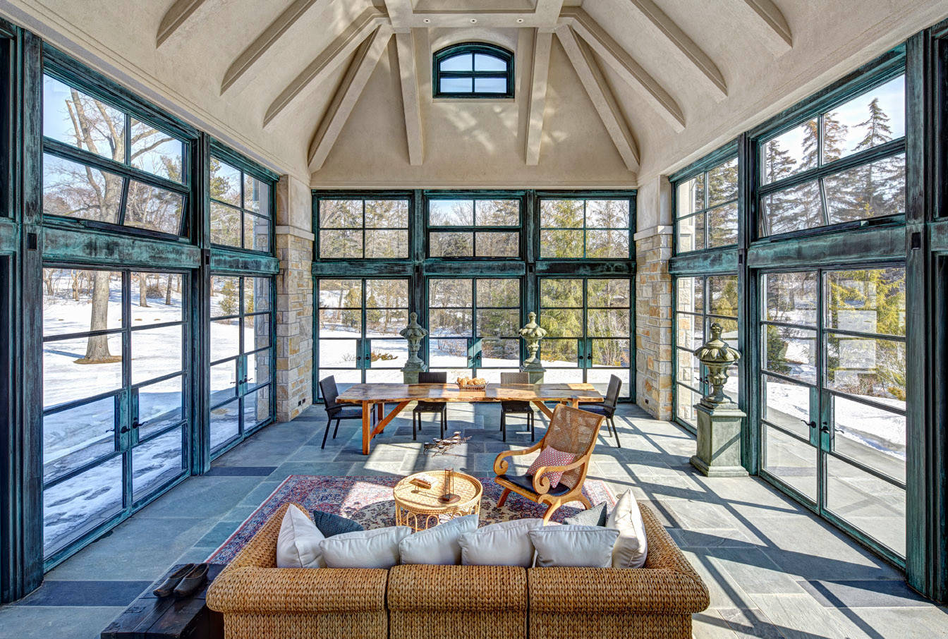 Vaulted solarium with patina-framed windows, woven furniture, slate floors, and snowy landscape views on all sides.