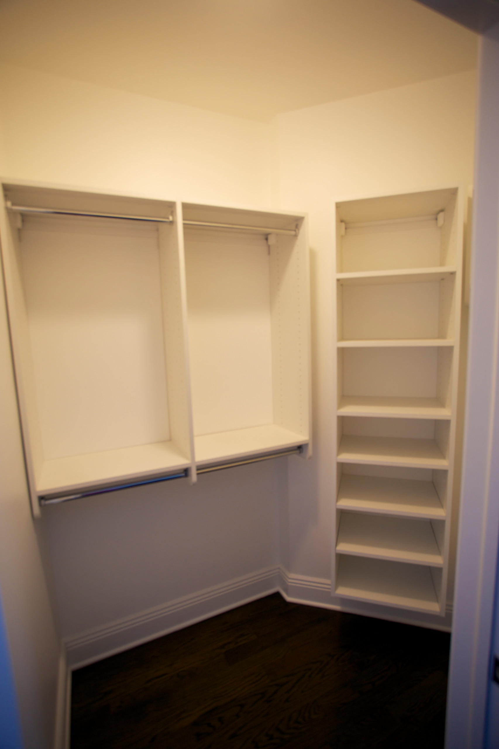 Corner walk-in closet featuring white built-in hanging sections, vertical shelving unit, and rich dark hardwood flooring.