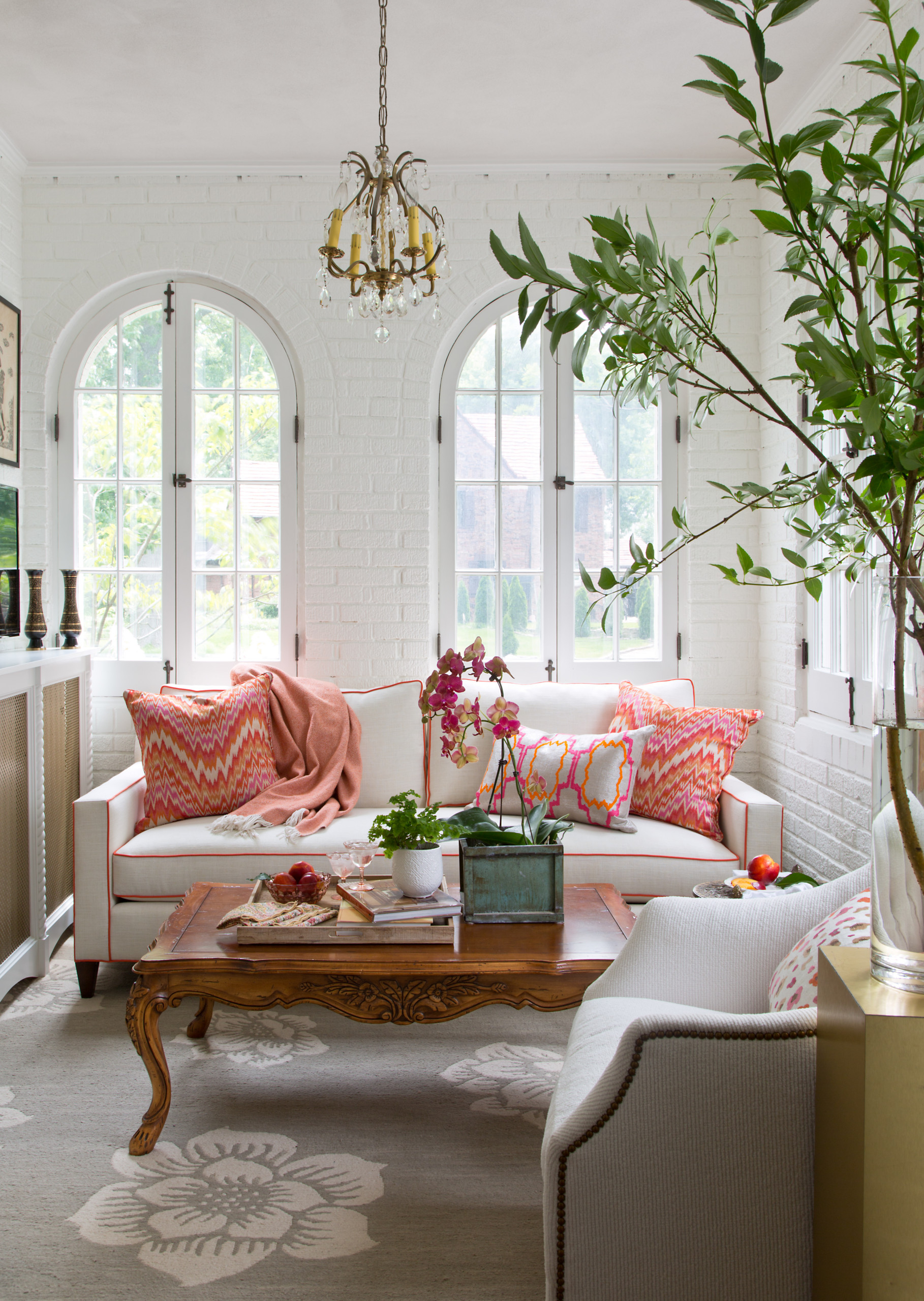 Bright living room with arched windows, white brick walls, coral-accented sofa, antique coffee table, and crystal chandelier.