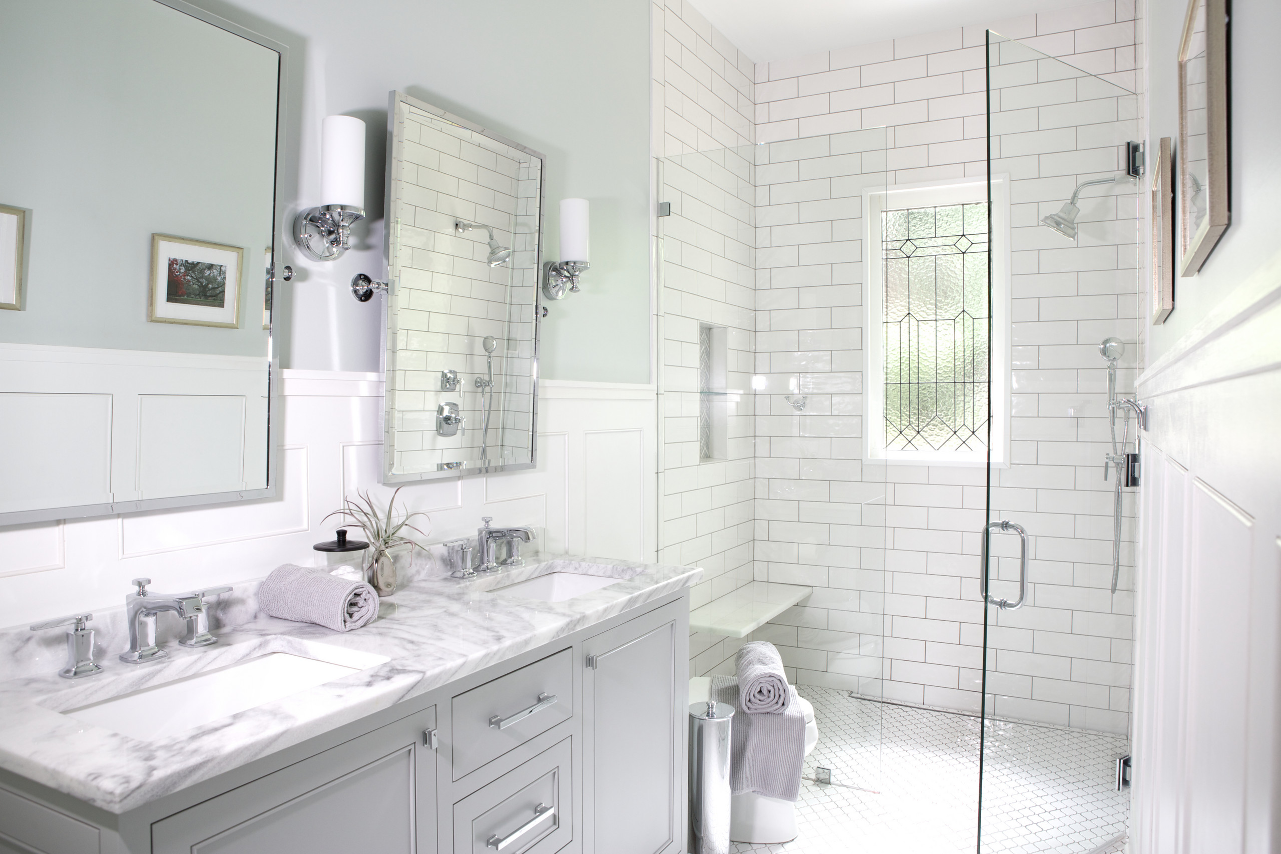 Bright white bathroom featuring marble countertops, subway tile walls, glass shower enclosure, and chrome fixtures with stained glass window.