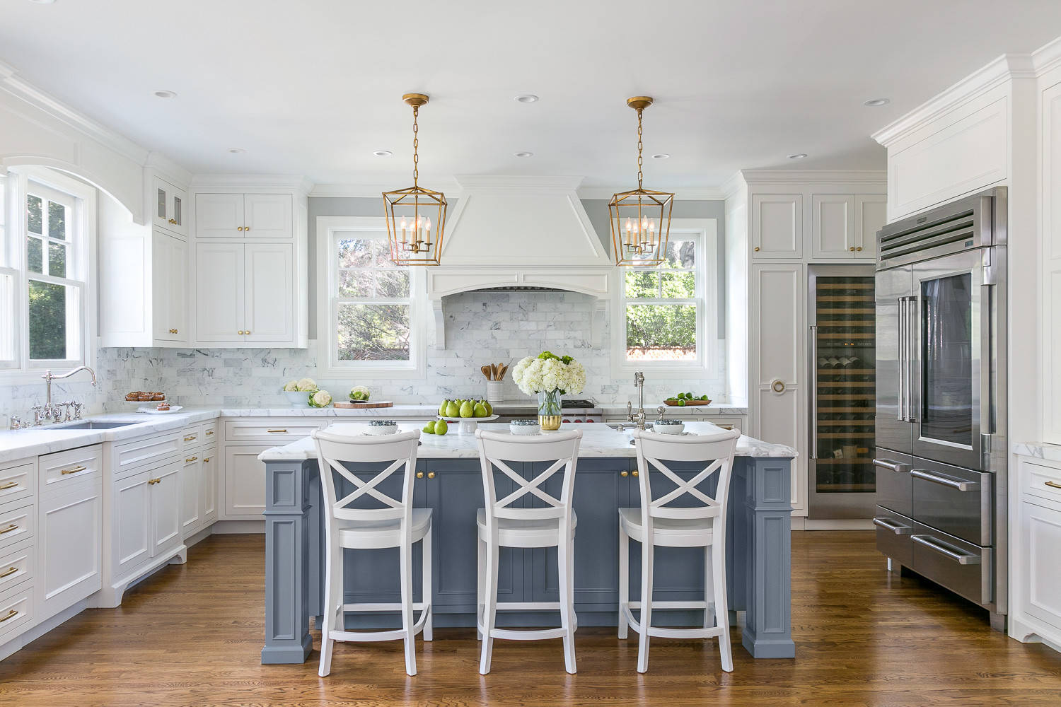 Bright white kitchen with marble backsplash, blue center island, brass lantern pendants, and white X-back barstools.
