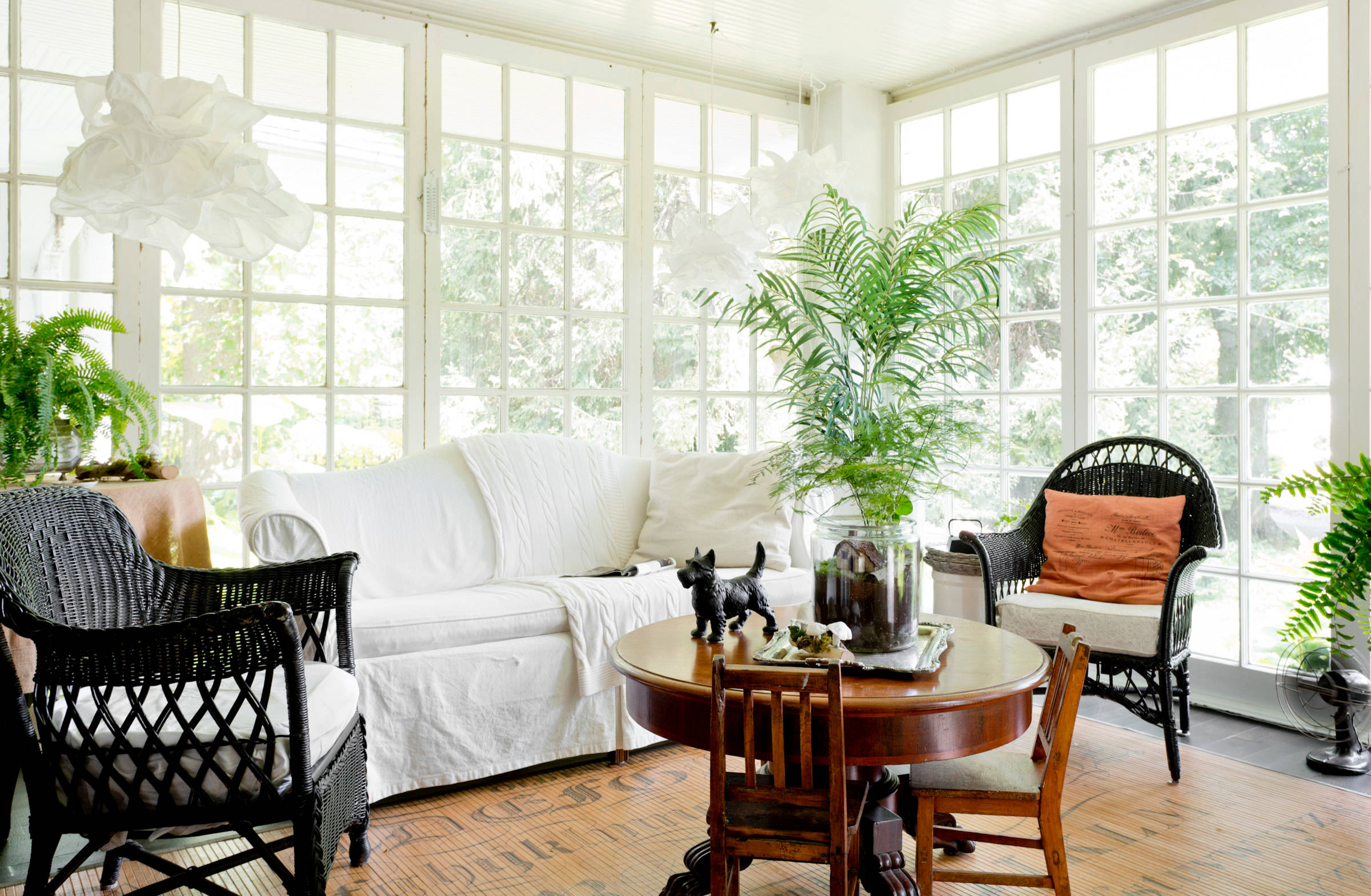 Light-filled solarium with white slipcovered sofa, black wicker chairs, round wooden table, and abundant greenery against gridded windows.