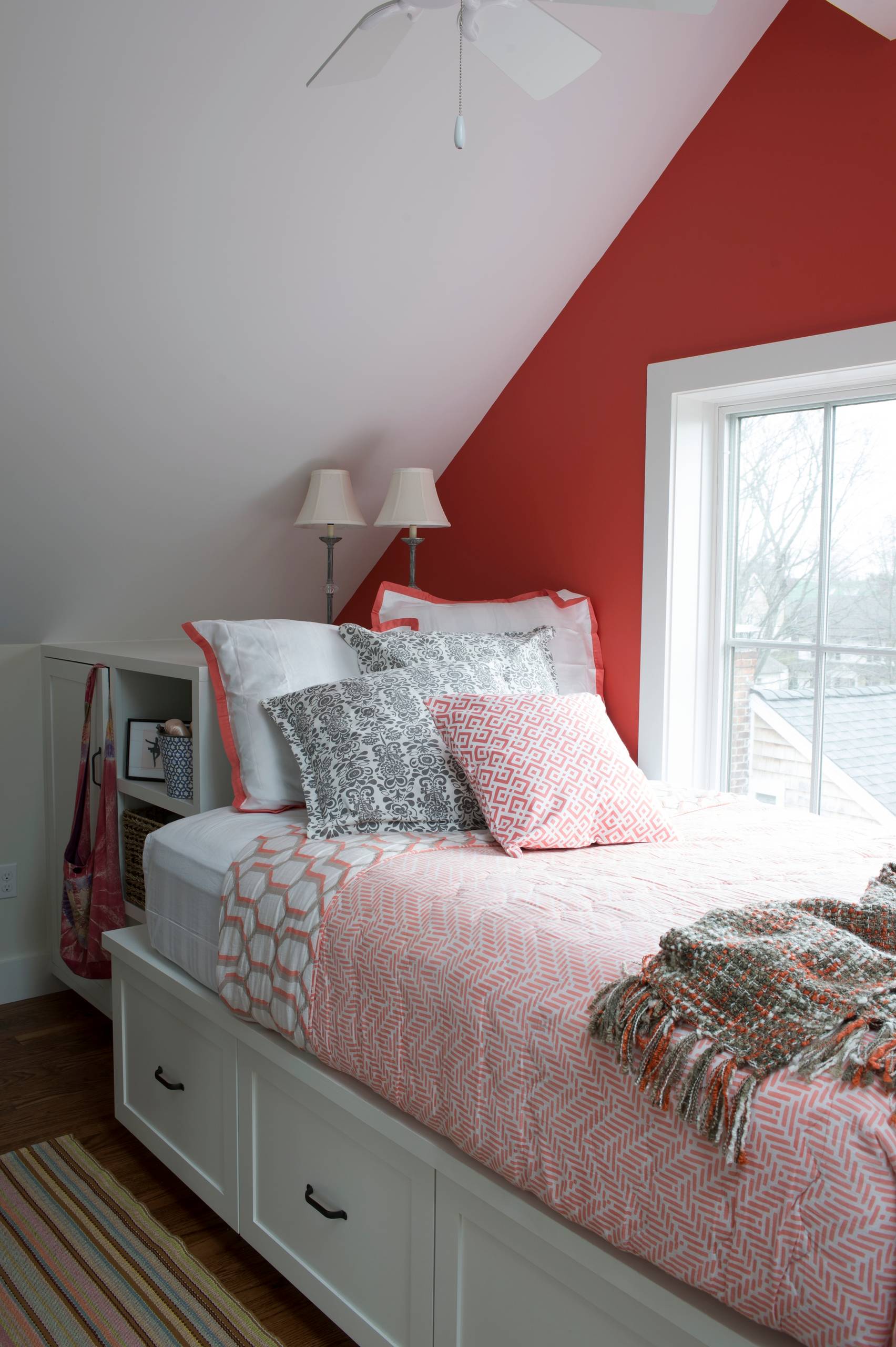 Attic bedroom with sloping white ceiling, bold red accent wall, built-in bed with storage drawers, and patterned bedding.