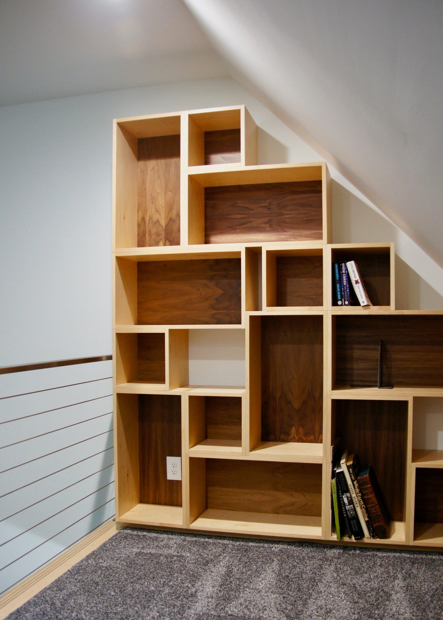 Modern light wood bookcase with dark wood back panels, built into an attic nook with sloped ceiling and light blue wall.