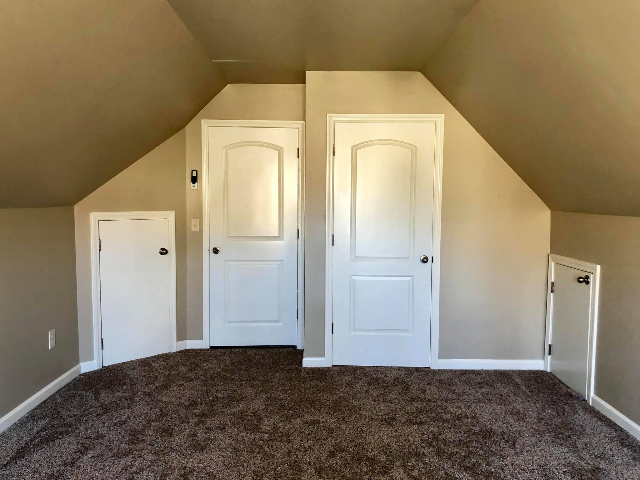 Empty attic room with sloped ceiling, beige walls, brown carpet, multiple white doors, and baseboards.