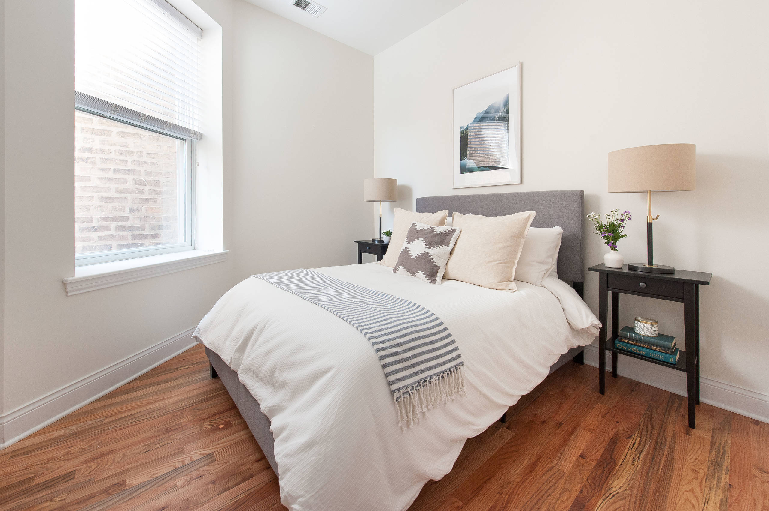 Bright bedroom with gray upholstered bed, white duvet, striped throw, dark wood nightstands, and hardwood floors.