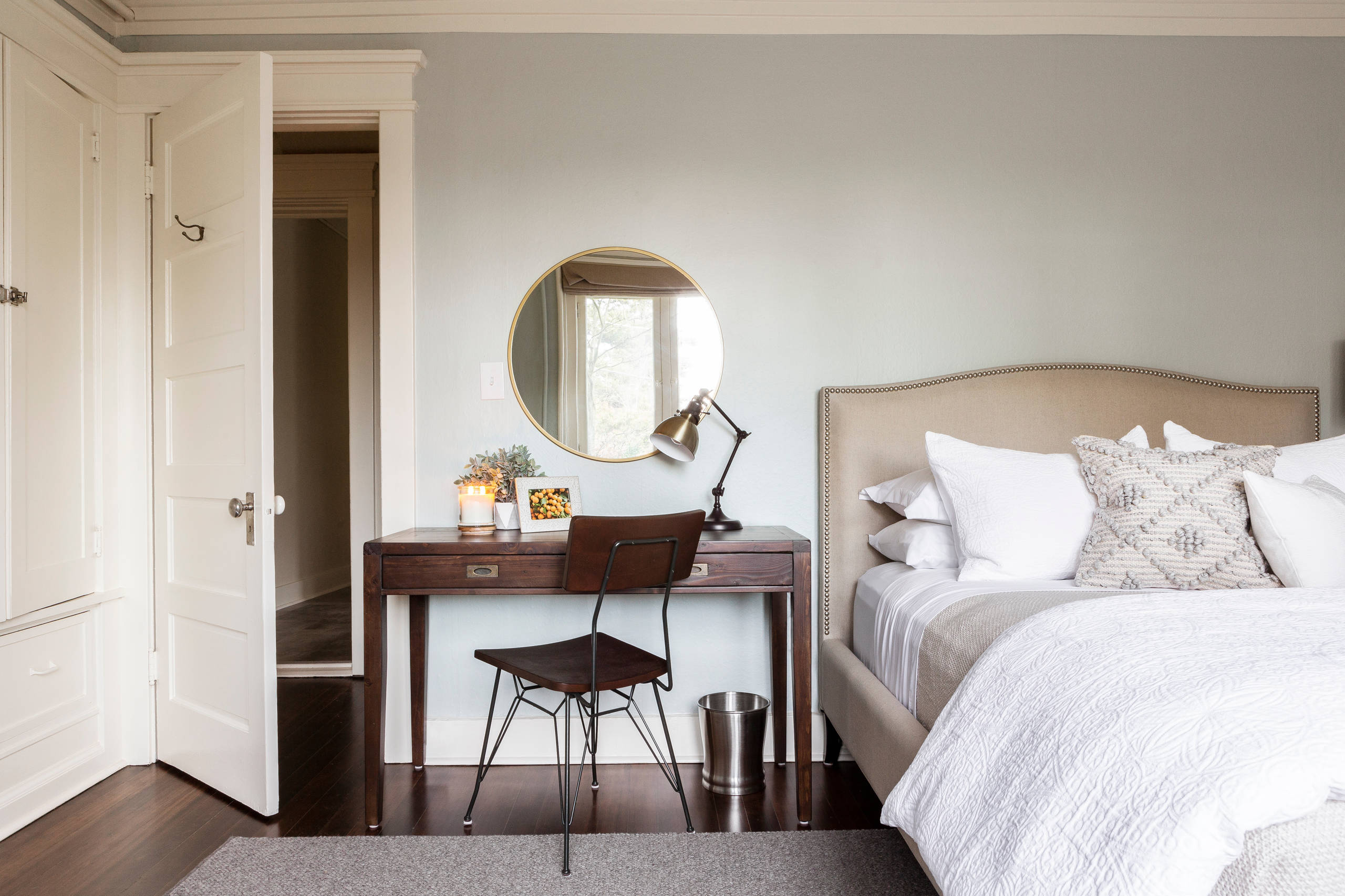 Neutral bedroom with upholstered bed, dark wood desk, round mirror, and white trim.