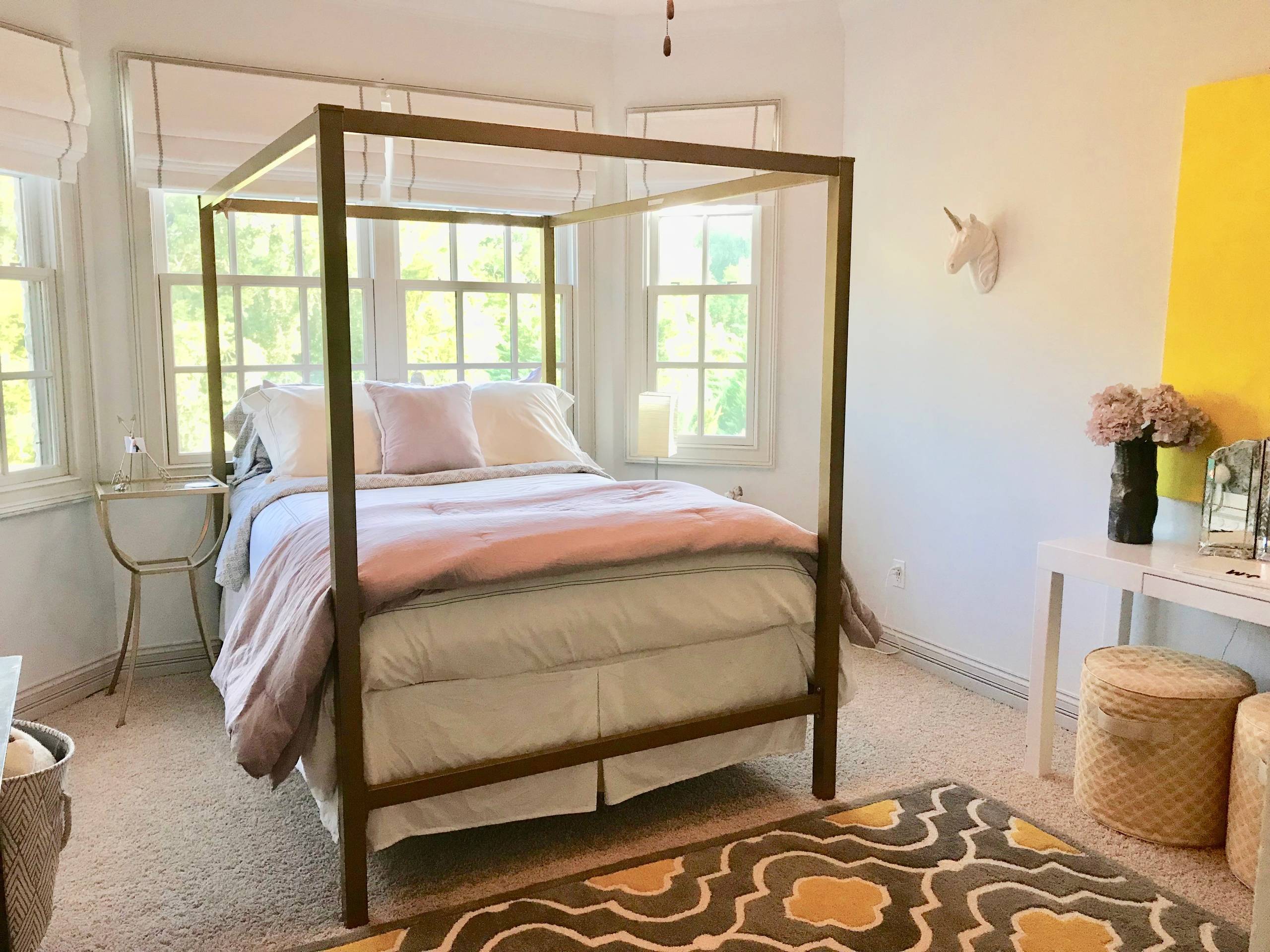 Youth bedroom with brass canopy bed, pink and white bedding, yellow accent wall, and patterned rug.