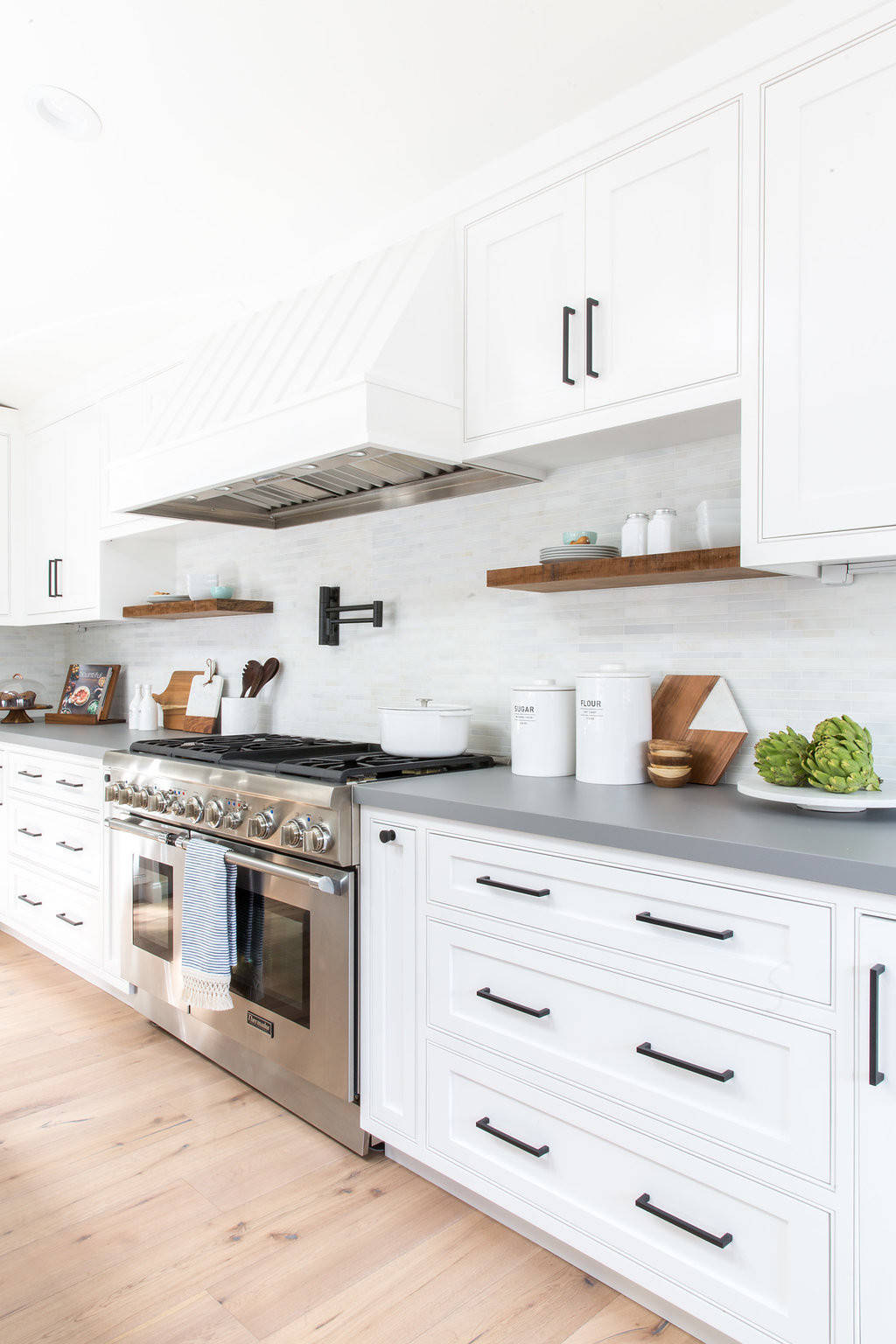 Bright modern kitchen with white shaker cabinets, stainless steel range, gray countertops, and wood open shelving.