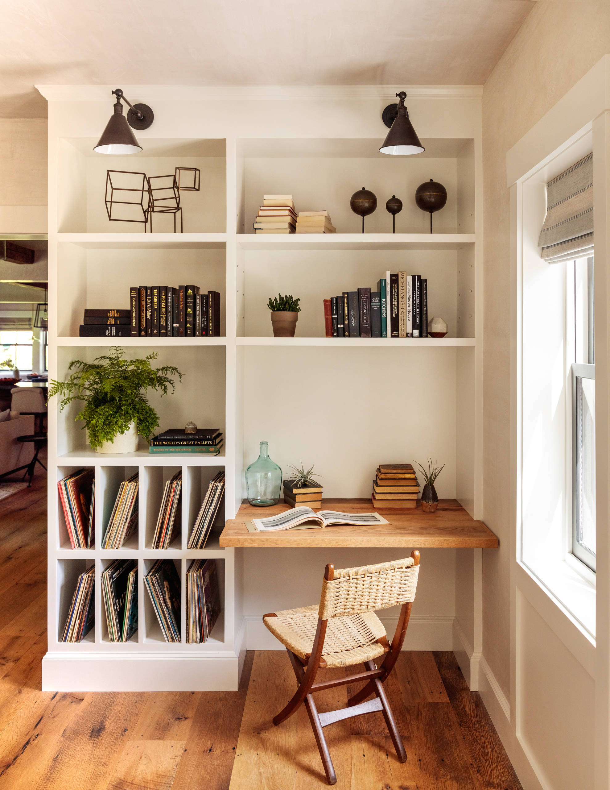 A cozy home office nook features white built-in shelving, a natural wood desk, and a woven chair on distressed hardwood floors.