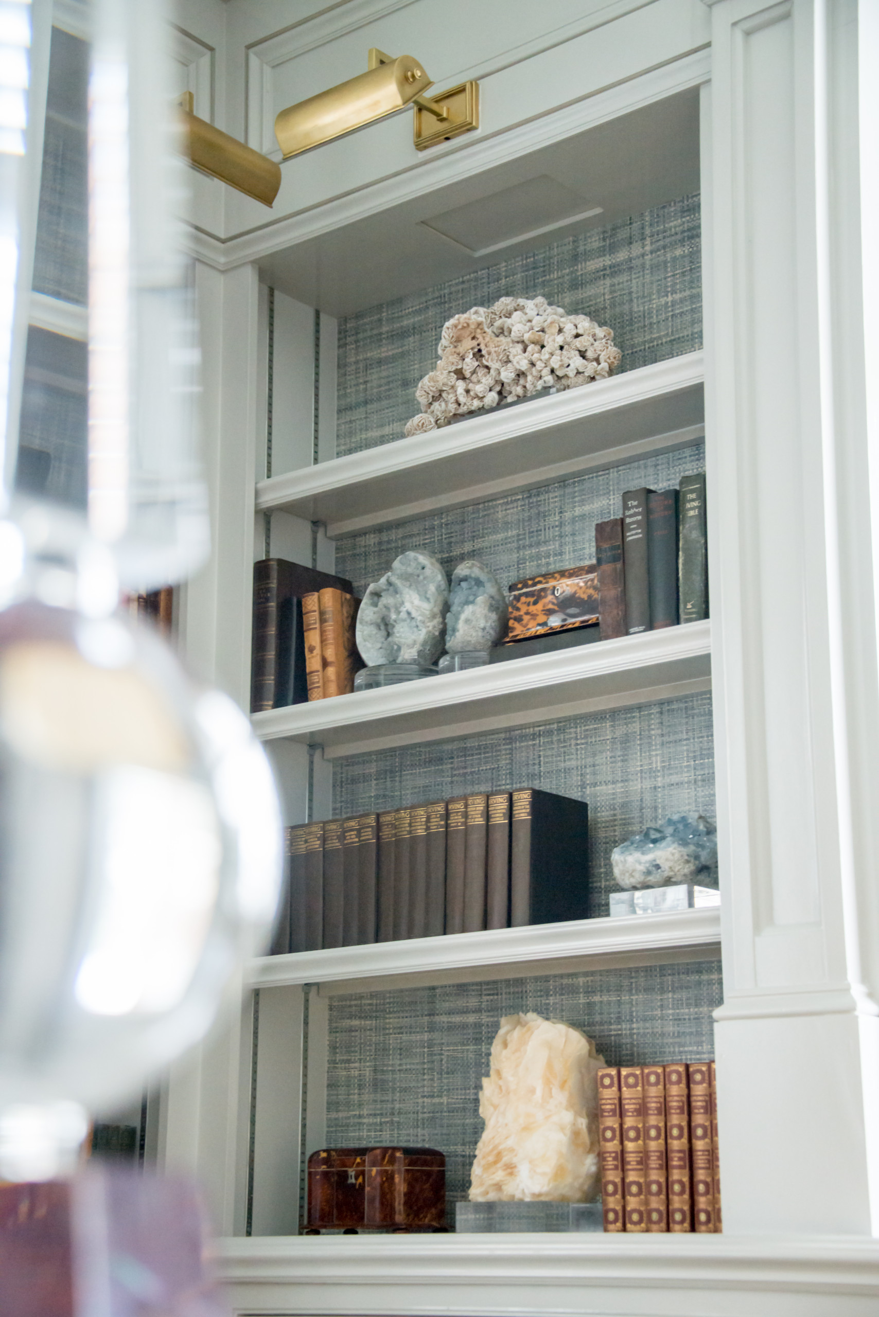 Custom white built-in bookshelf with blue textured wallpaper, brass library lights, and various books and geological specimens.