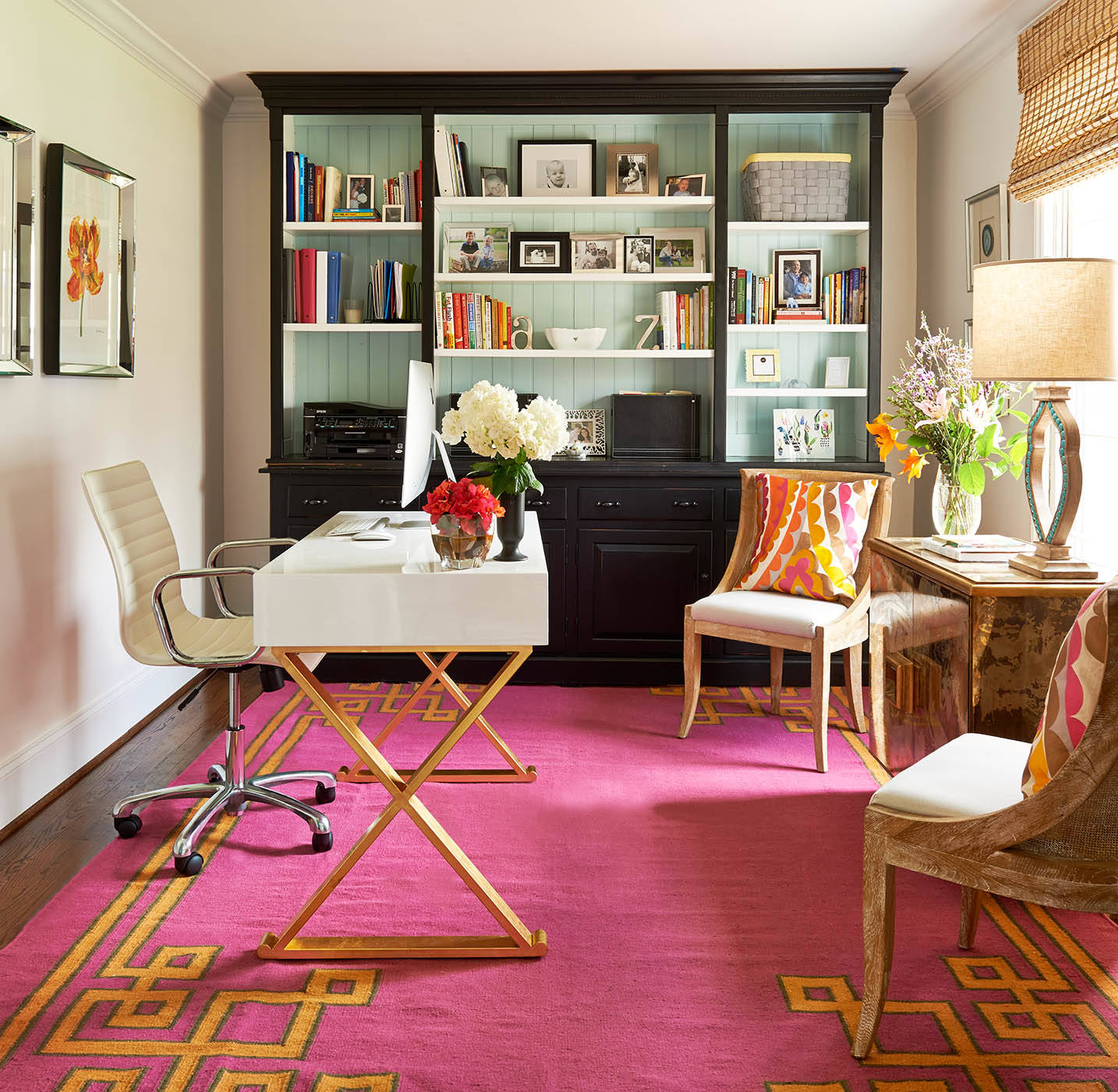 Vibrant home office with a fuchsia rug, black built-in shelving, white modern desk, and rattan chairs.