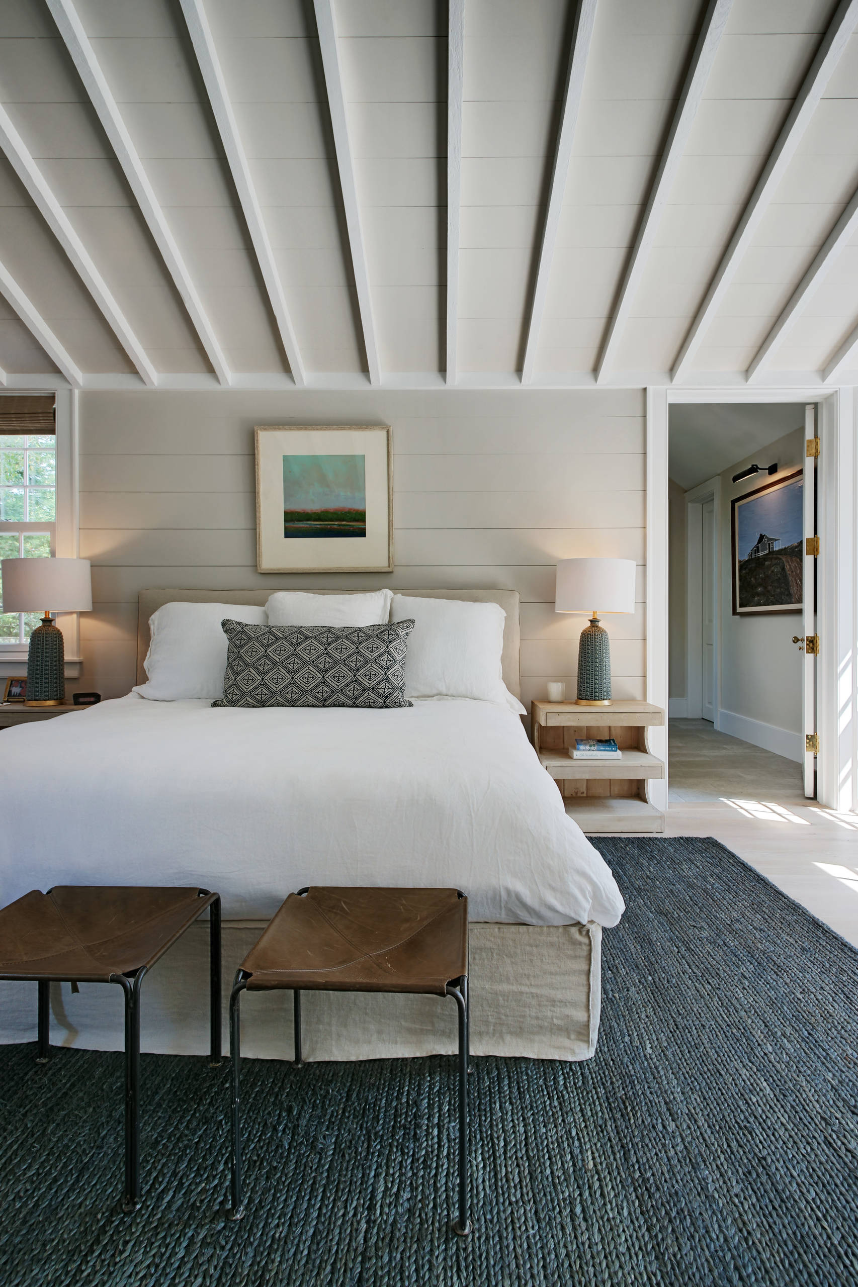 Bright bedroom with exposed white rafter ceiling, shiplap walls, blue woven rug, and light wood furniture.