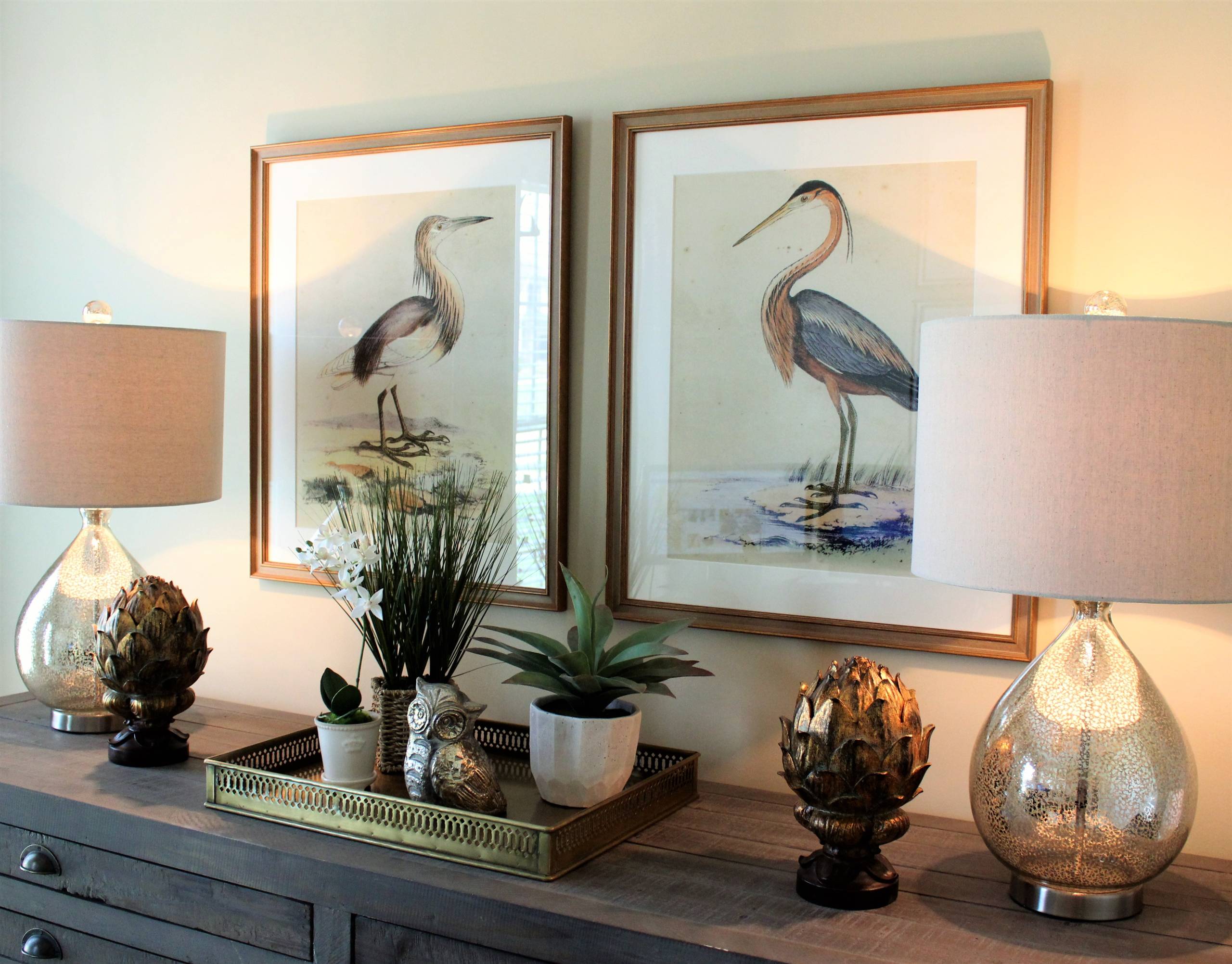 Weathered gray console with mercury glass lamps, gold artichoke finials, brass tray, and two framed bird prints.