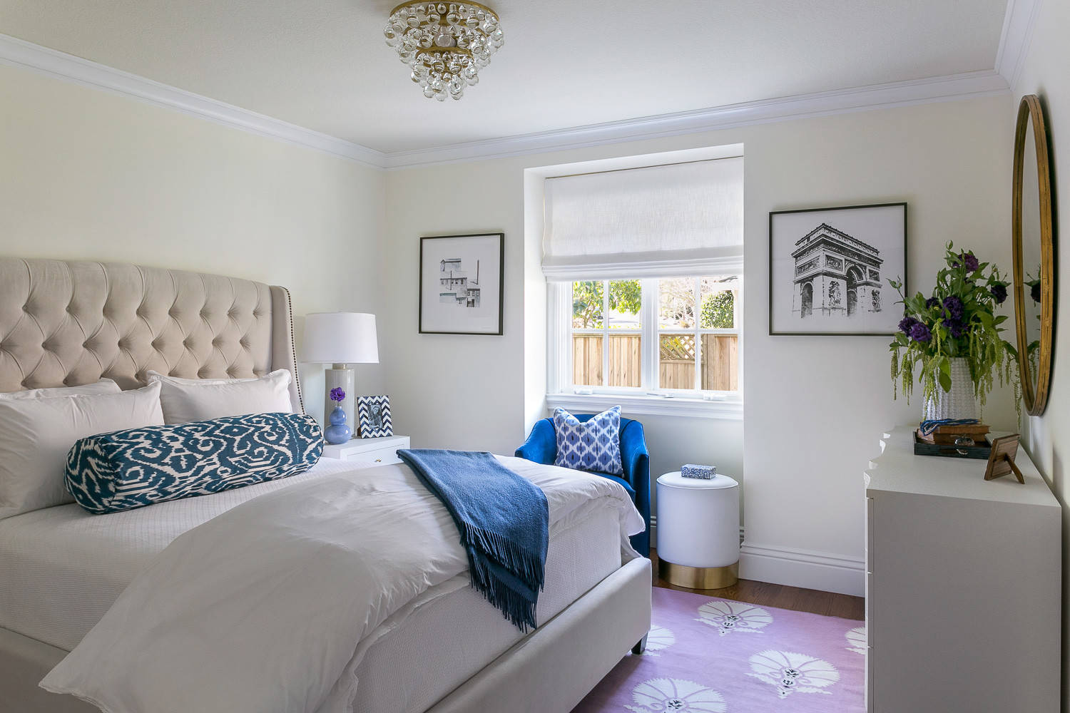 Bright bedroom with tufted neutral bed, purple floral rug, blue velvet chair, and gold chandelier.