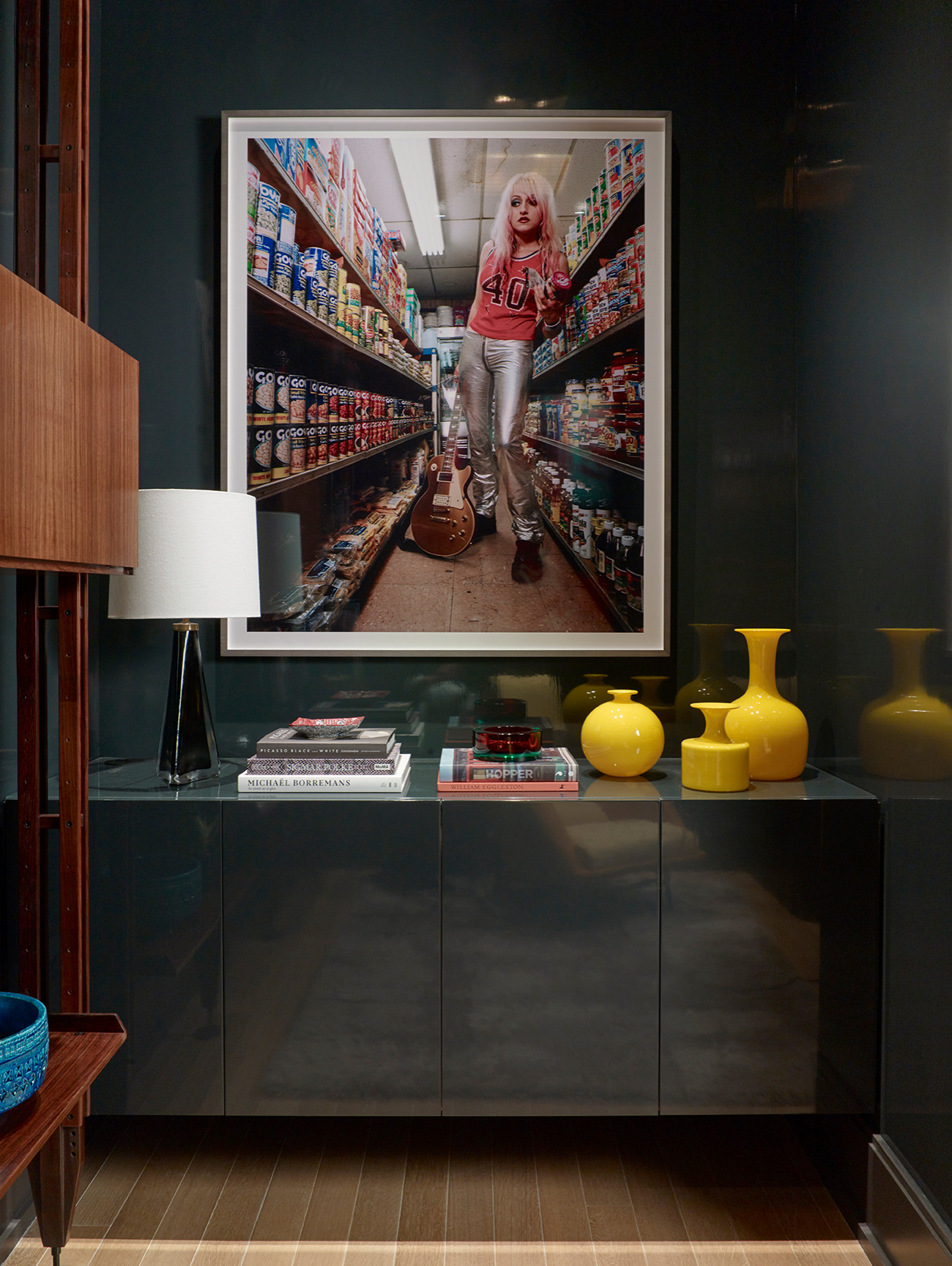 High-gloss teal console cabinet with vibrant yellow vases, a white lamp, and striking framed artwork against a dark wall.