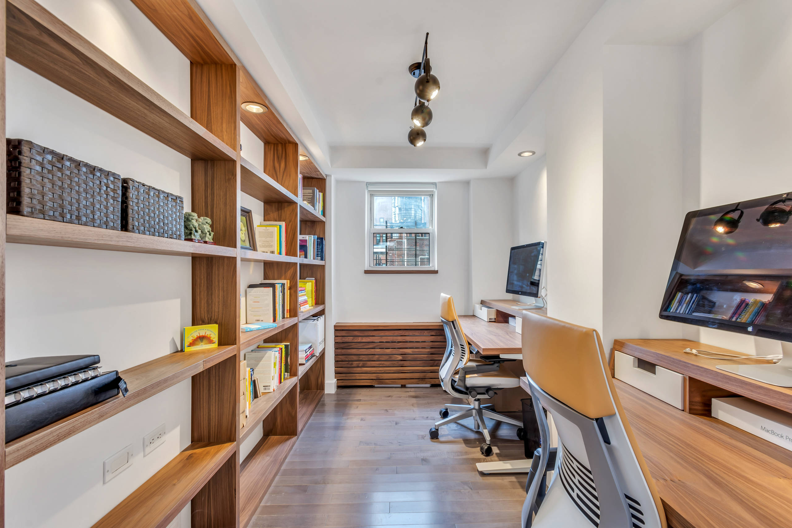 Sleek home office with warm wood built-in shelving, dual desks, and light wood flooring, bathed in soft recessed lighting.