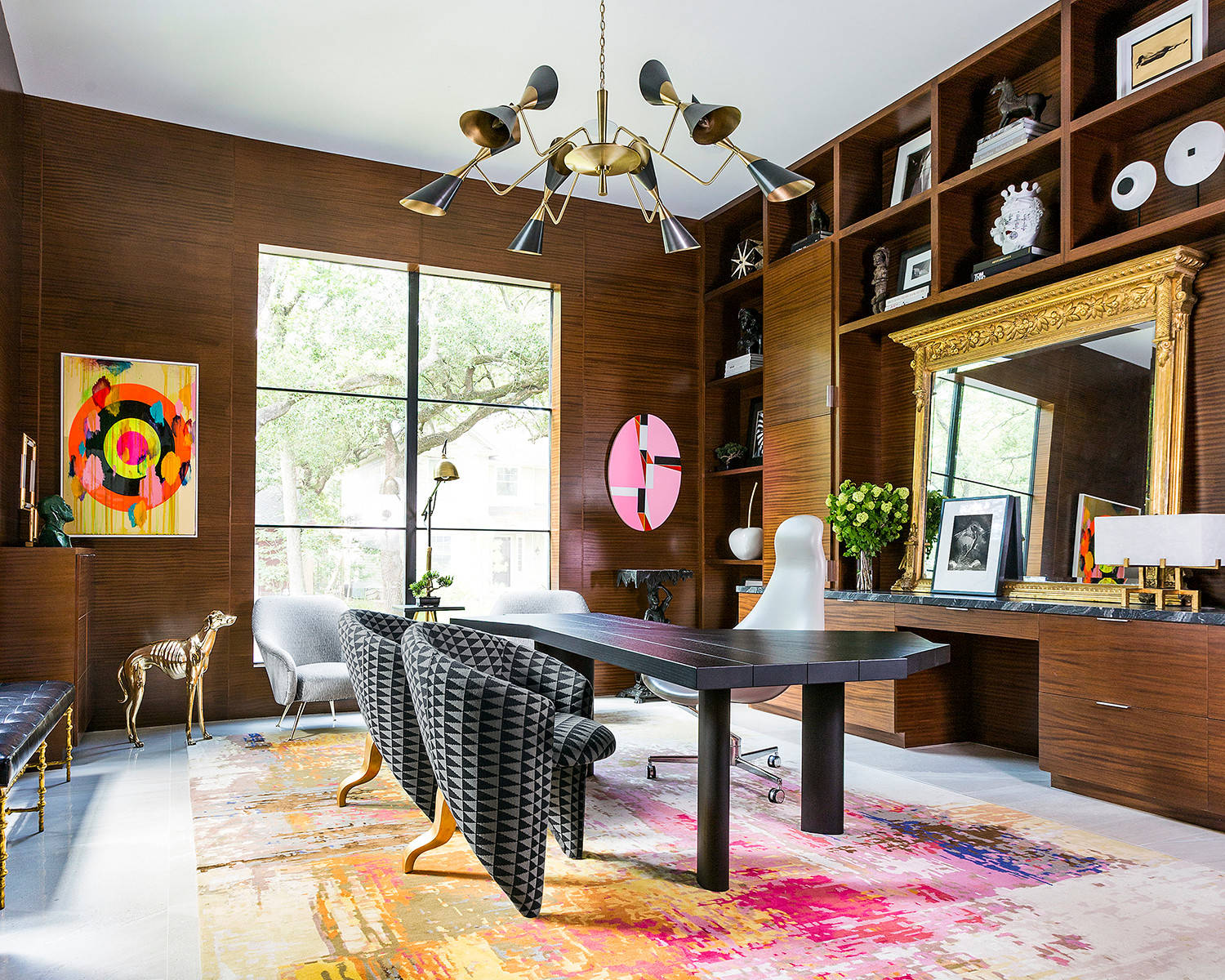 Warm wood-paneled office with vibrant abstract art, a black desk, patterned chairs, and a brass chandelier.