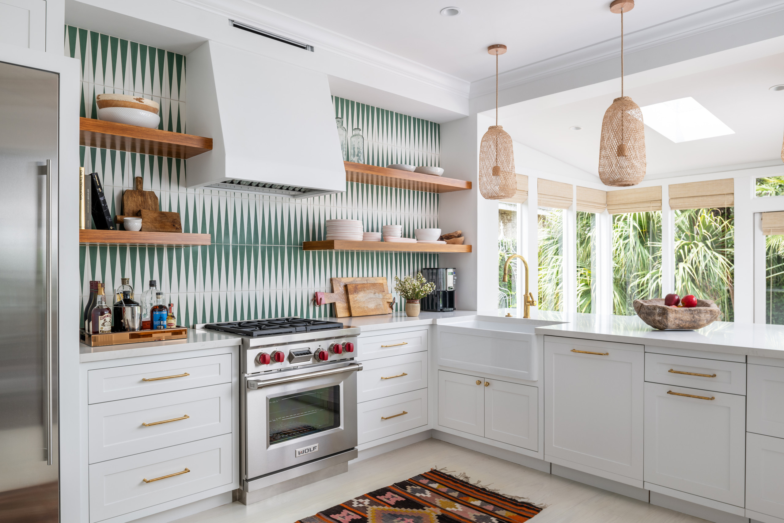 Bright kitchen with white cabinets, green geometric backsplash, natural wood shelves, and woven pendants over a window.