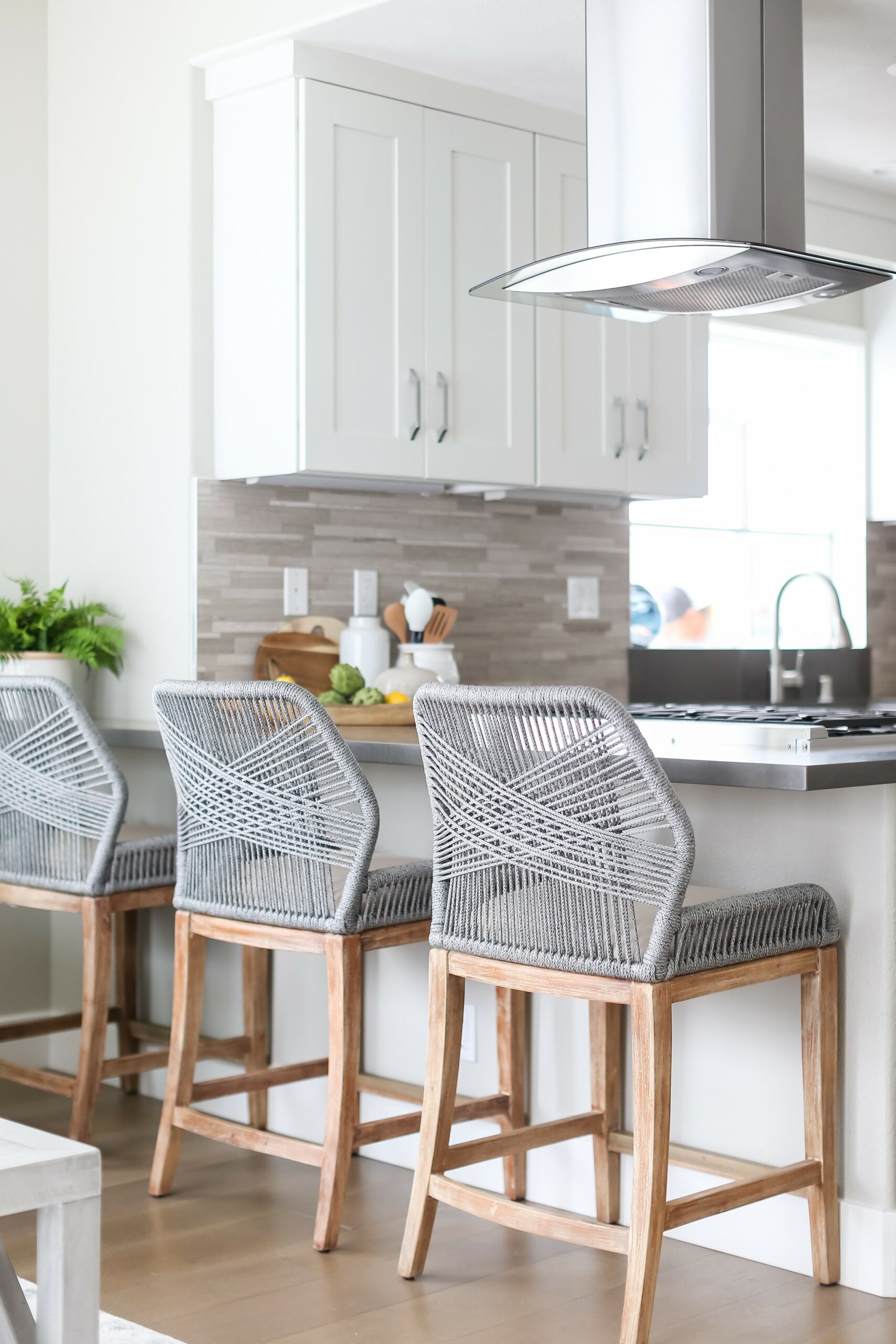Bright kitchen featuring sleek white cabinetry, gray rope barstools, and a textured rectangular tile backsplash.