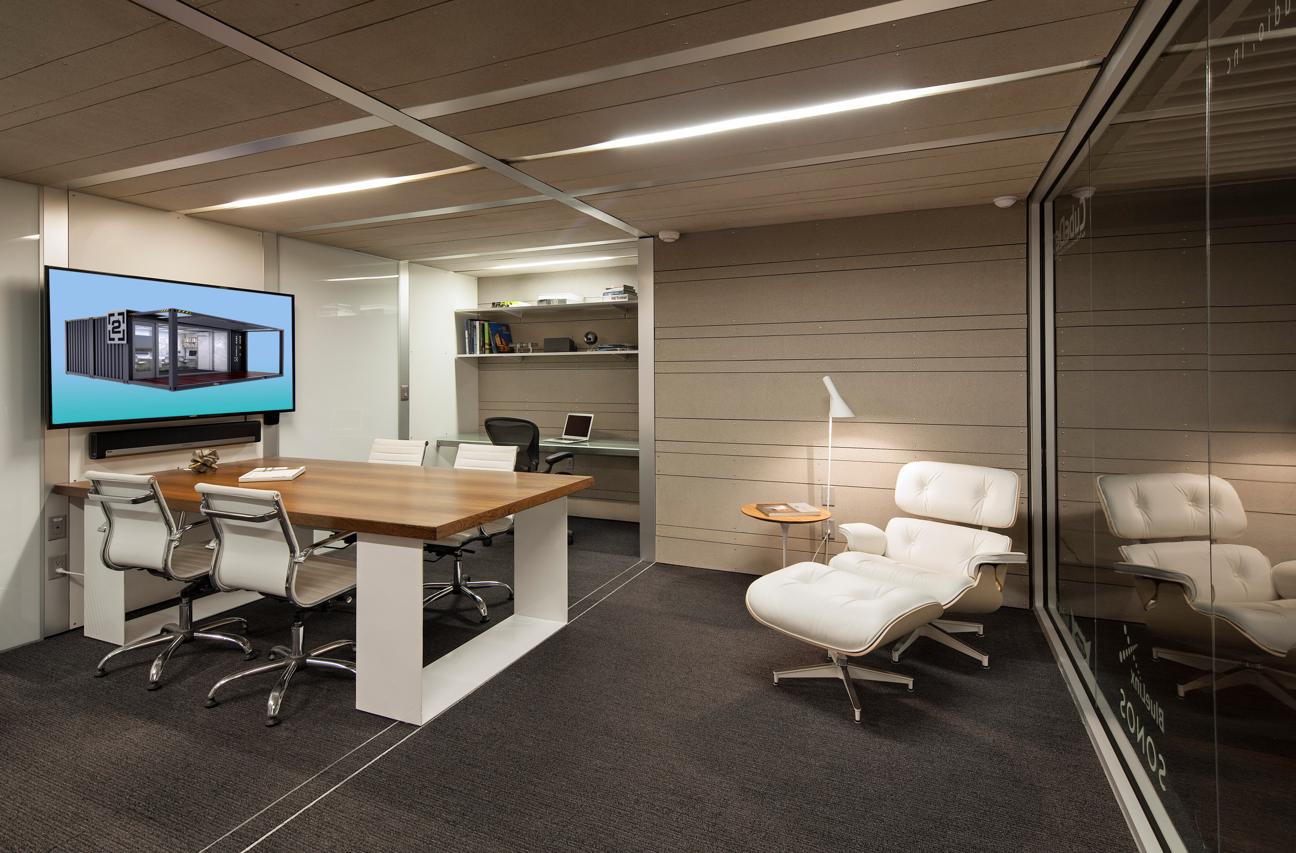 A modern office space featuring a large wooden conference table, white chairs, and a cozy lounge area with a white leather armchair.