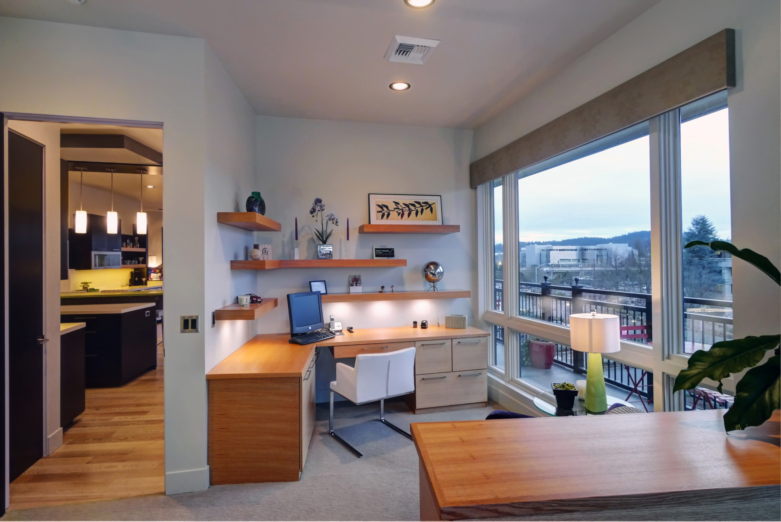 Warm wood desk and floating shelves in a contemporary home office with large windows overlooking a city.