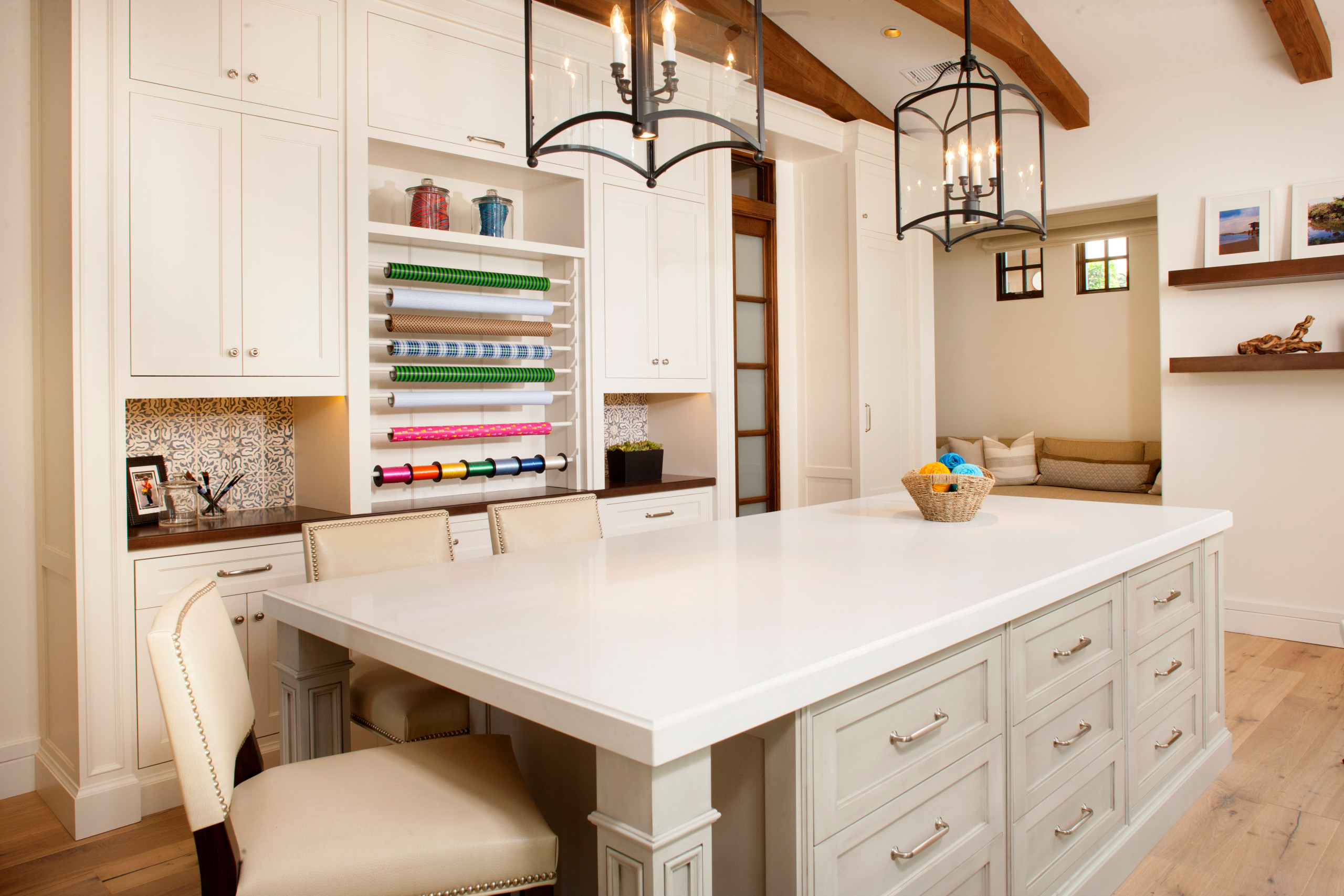 Bright craft room with white cabinets, large central island, and a cozy reading nook beyond.