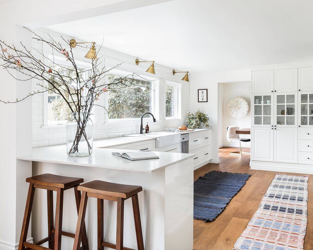 Bright farmhouse kitchen with white subway tile, brass sconces, wood stools, and a large white cabinet system.