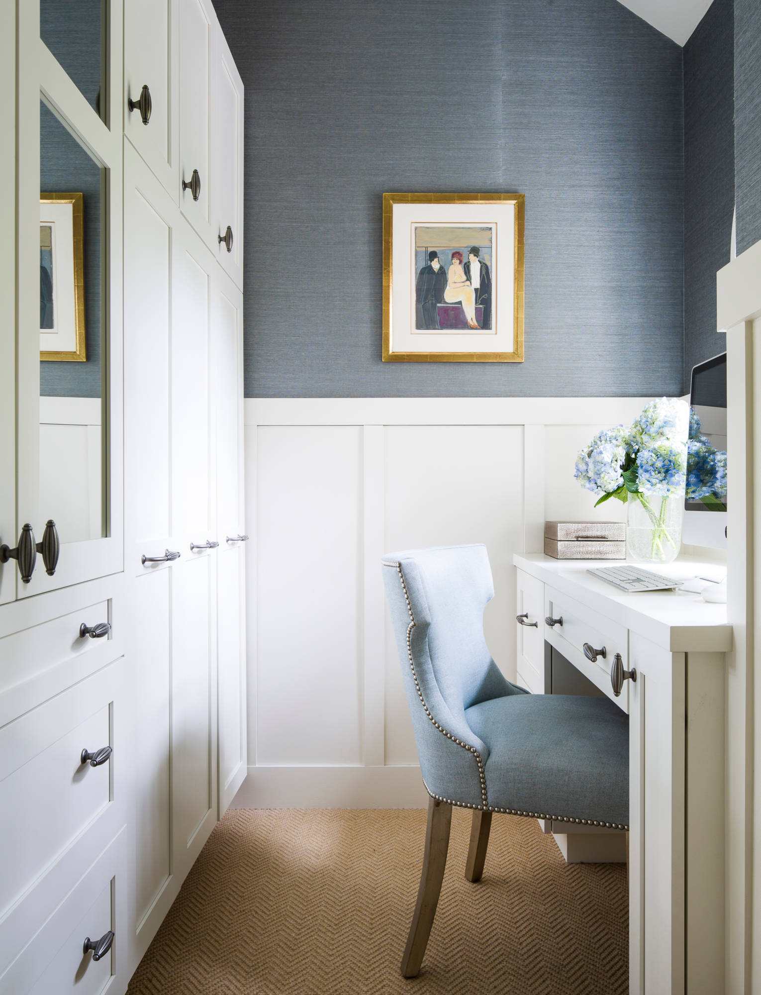 Compact home office with blue-gray grasscloth walls, white wainscoting, built-in storage, and a light blue upholstered chair.