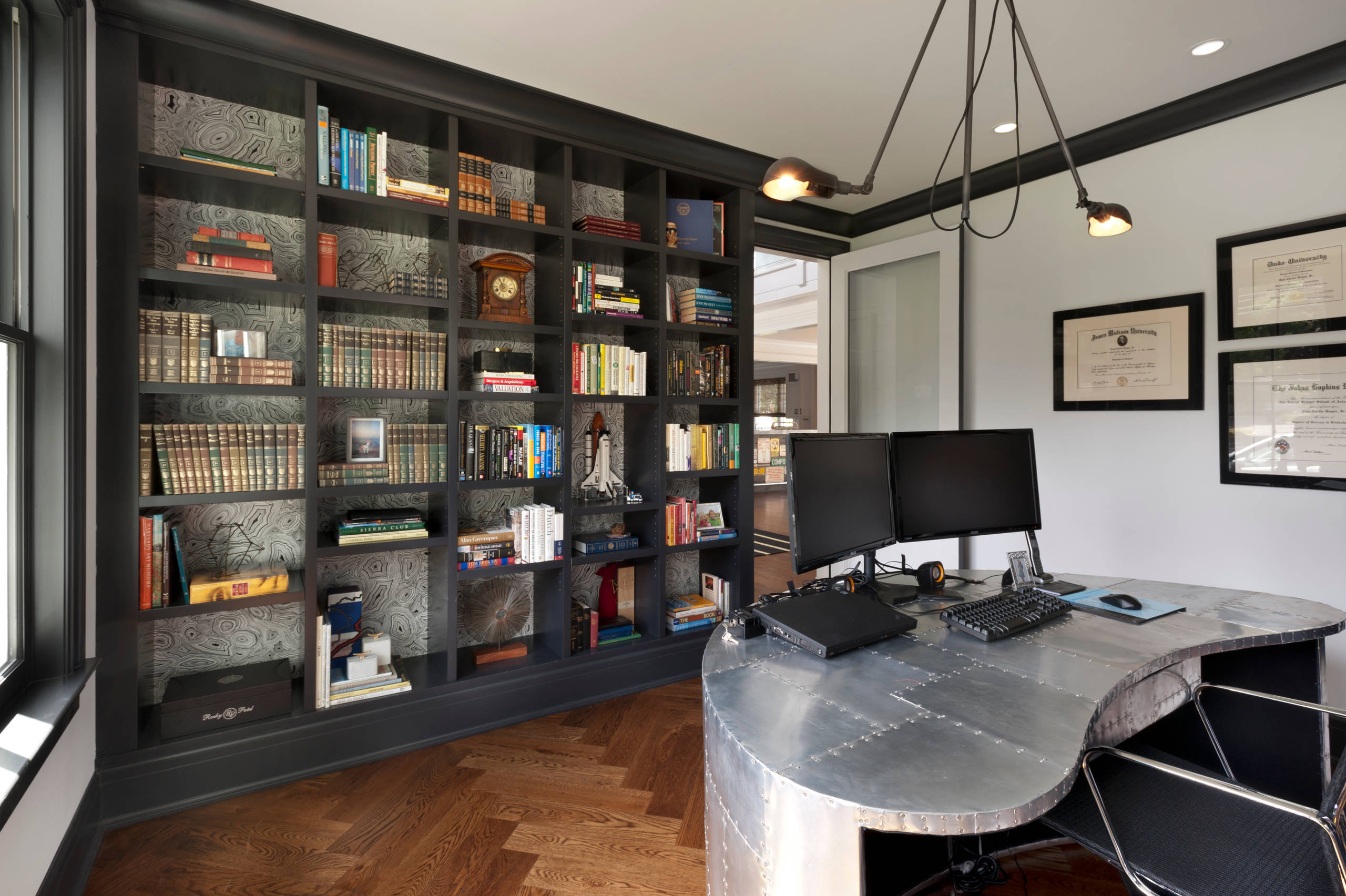 Home office with dark built-in bookcase, patterned wallpaper, metal desk, and herringbone wood floor.