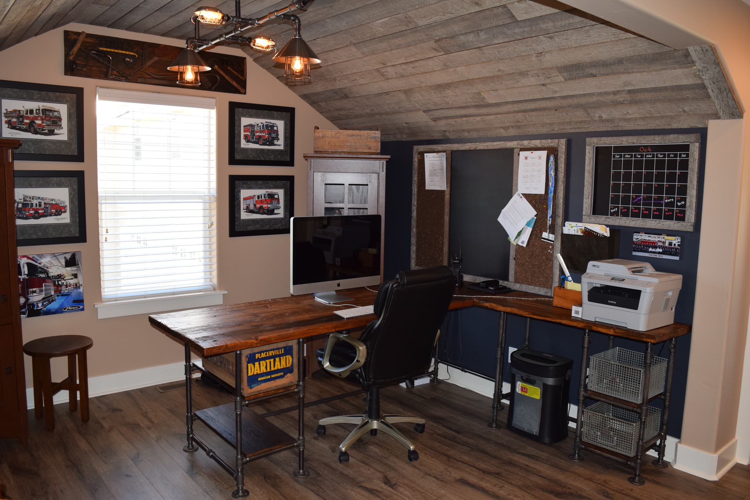 A rustic industrial home office with a dark wood desk, pipe shelving, distressed wood ceiling, and fire truck art.