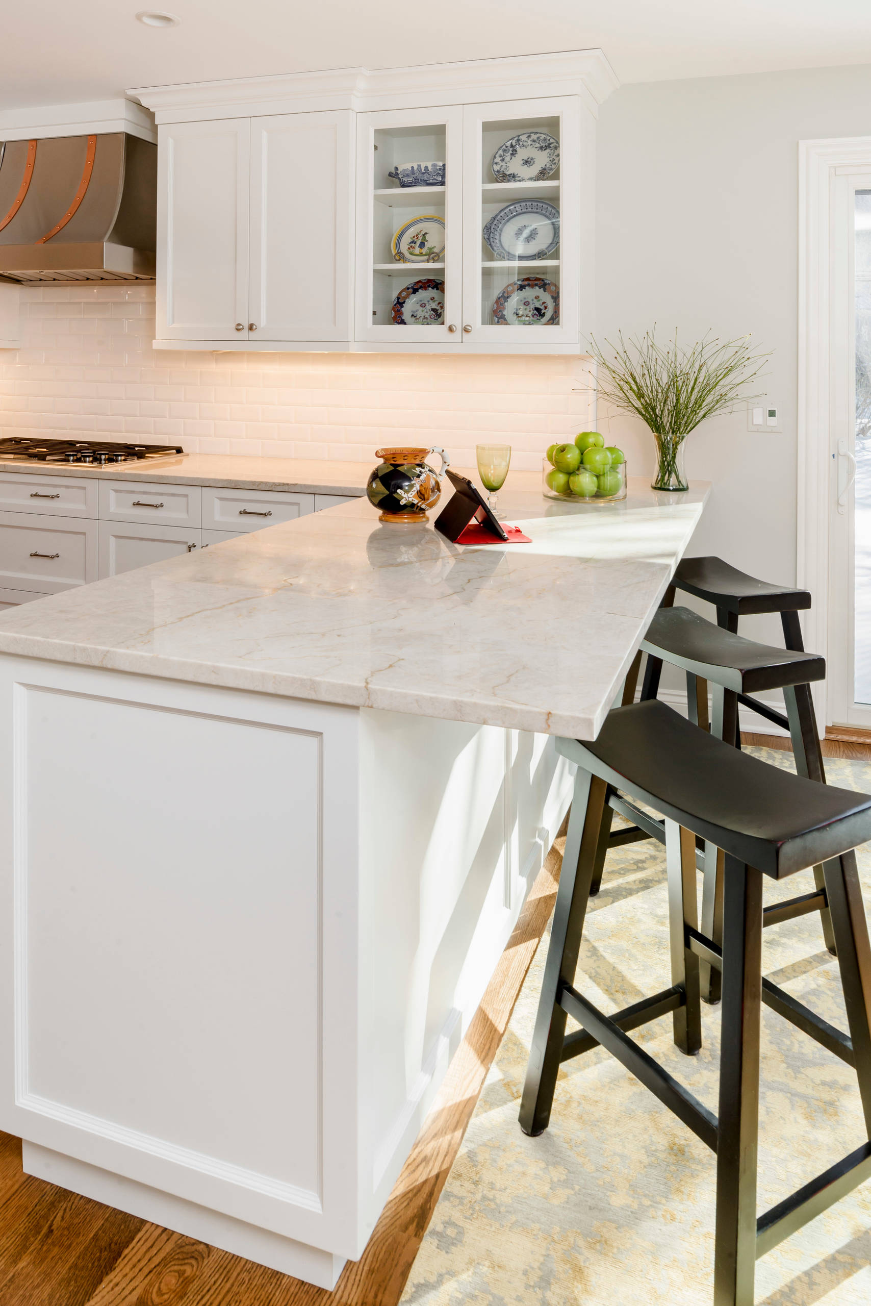 Bright kitchen with white shaker cabinets, subway tile backsplash, marble island, dark stools, and copper range hood detail.