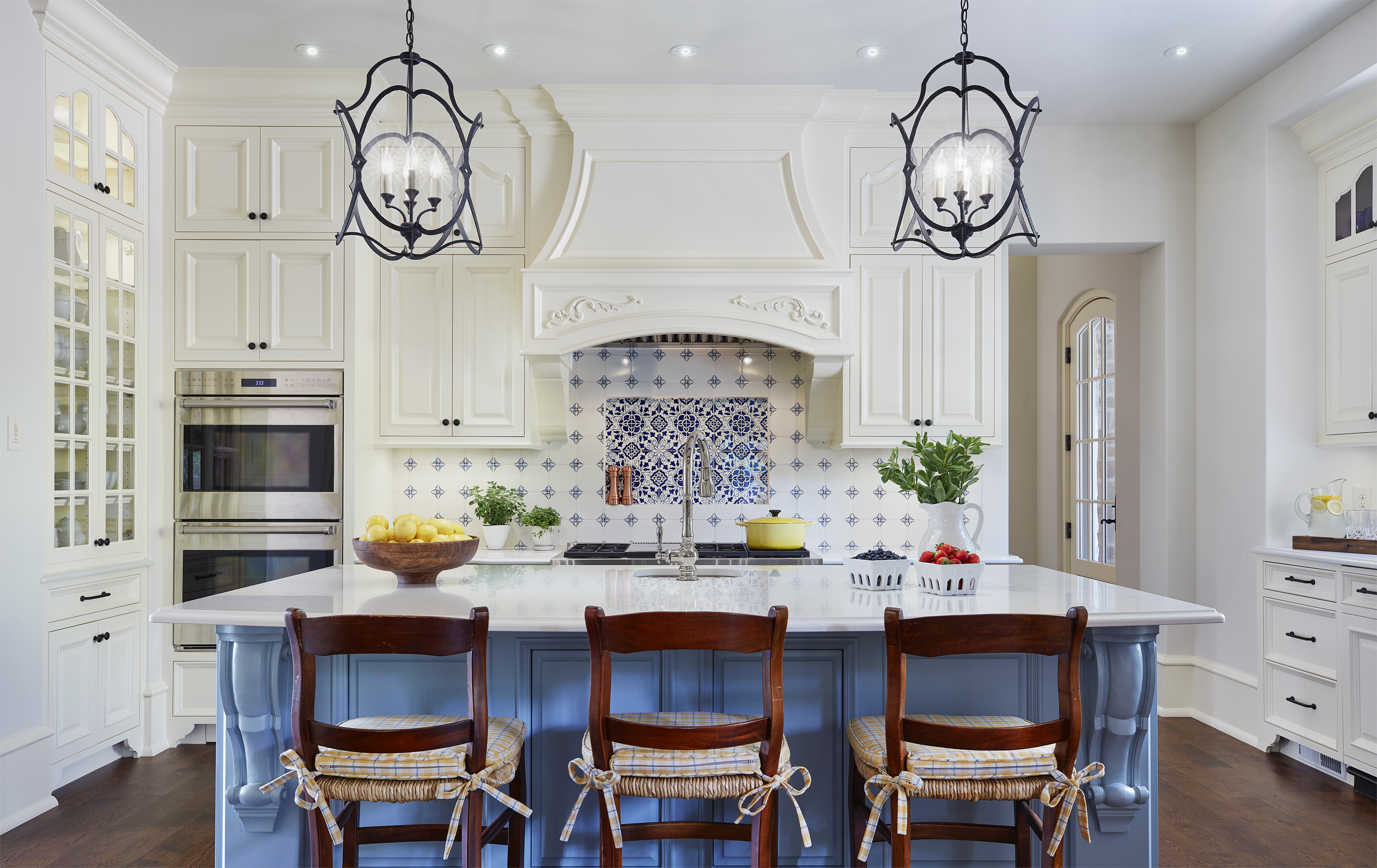 White kitchen with blue tiled backsplash, large island with gray-blue base, dark wood chairs, and wrought iron pendants.