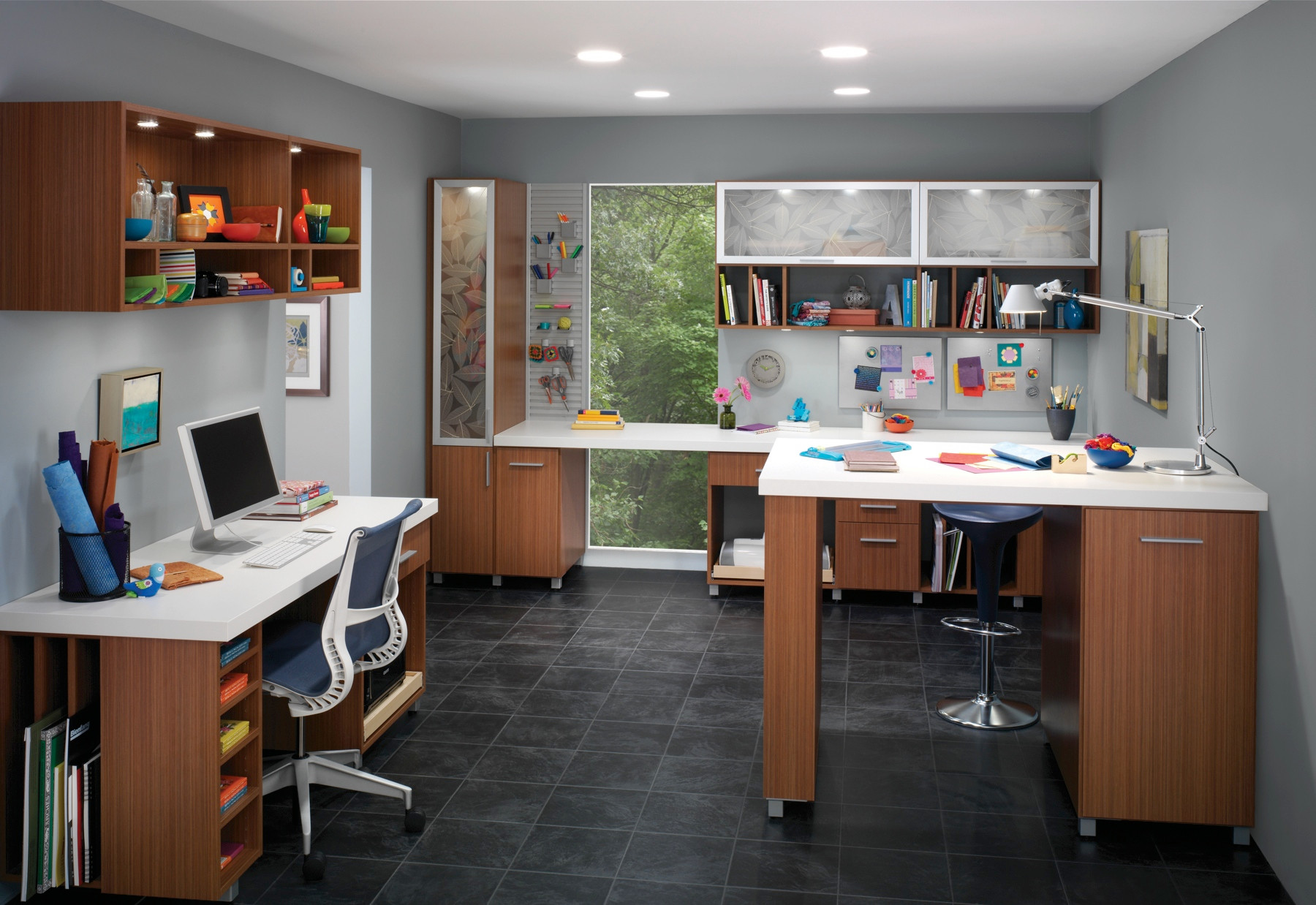 A creative studio with warm wood cabinetry, white countertops, ergonomic chairs, and a large window overlooking greenery.